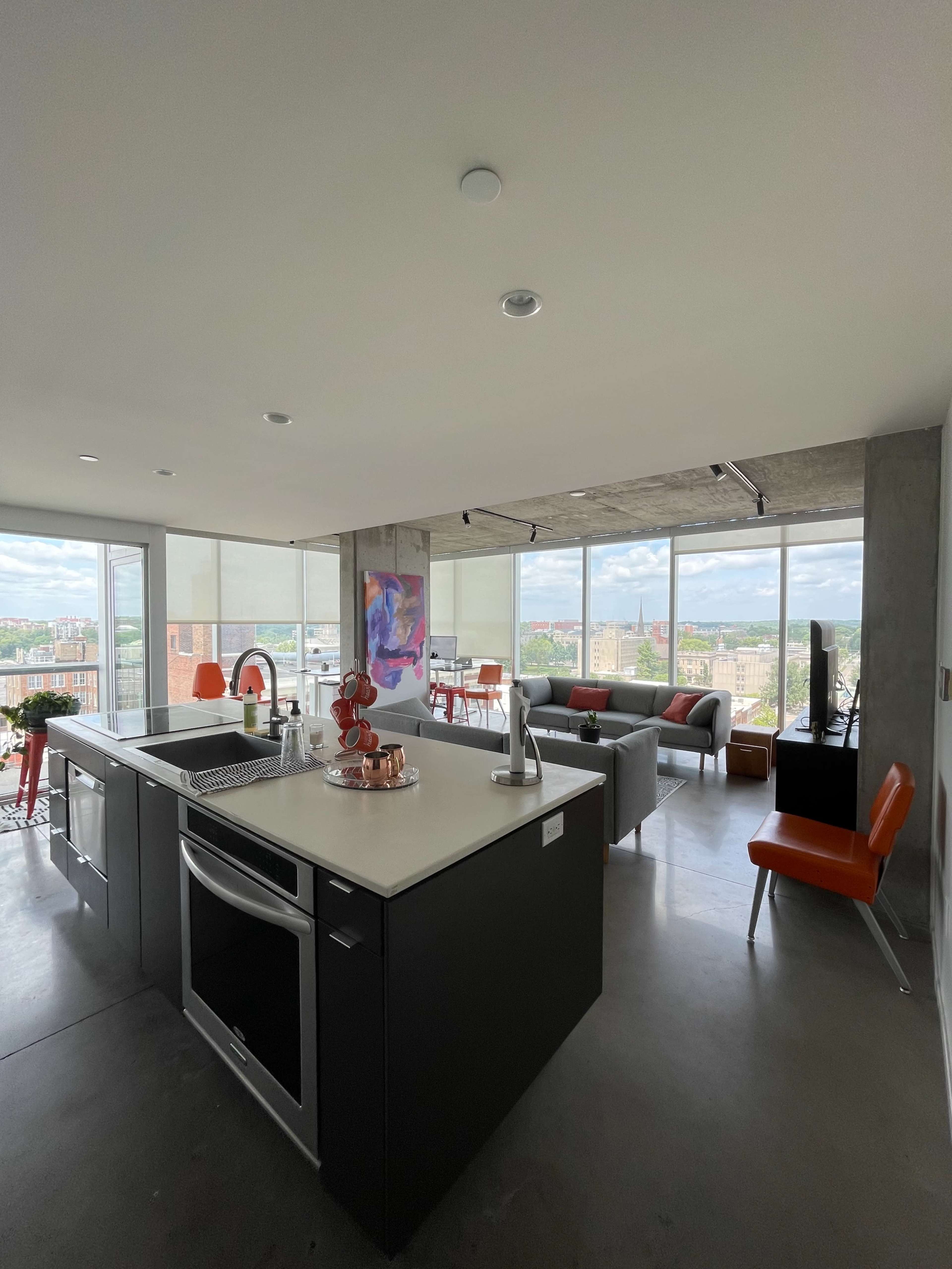 A modern, open-concept apartment features a kitchen with a large island and floor-to-ceiling windows that offer a view of the city.