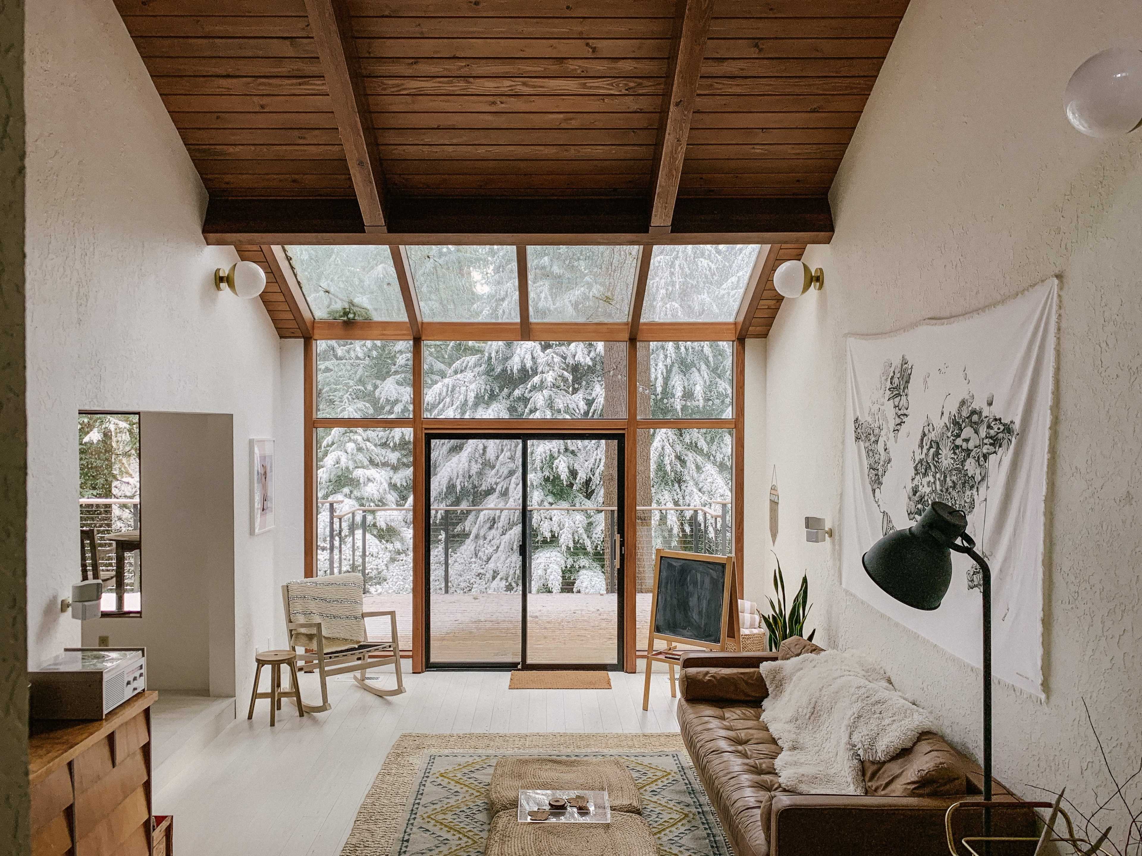 A cozy living room with large windows revealing a snowy landscape outside, complemented by wooden beams and minimalist furniture.