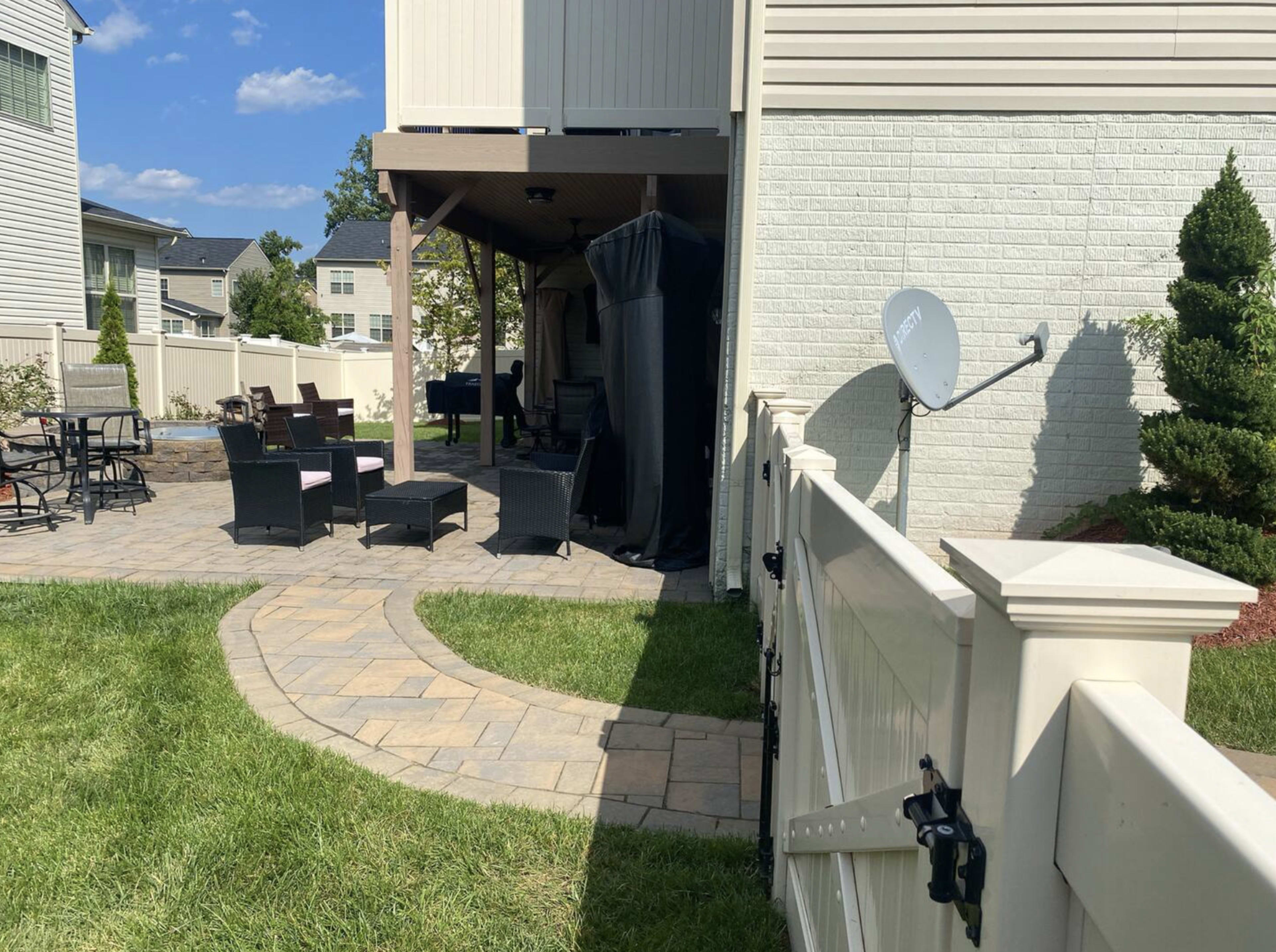 The image shows a landscaped backyard with a stone pathway leading to a patio area, surrounded by a fence and shaded by an upper deck.