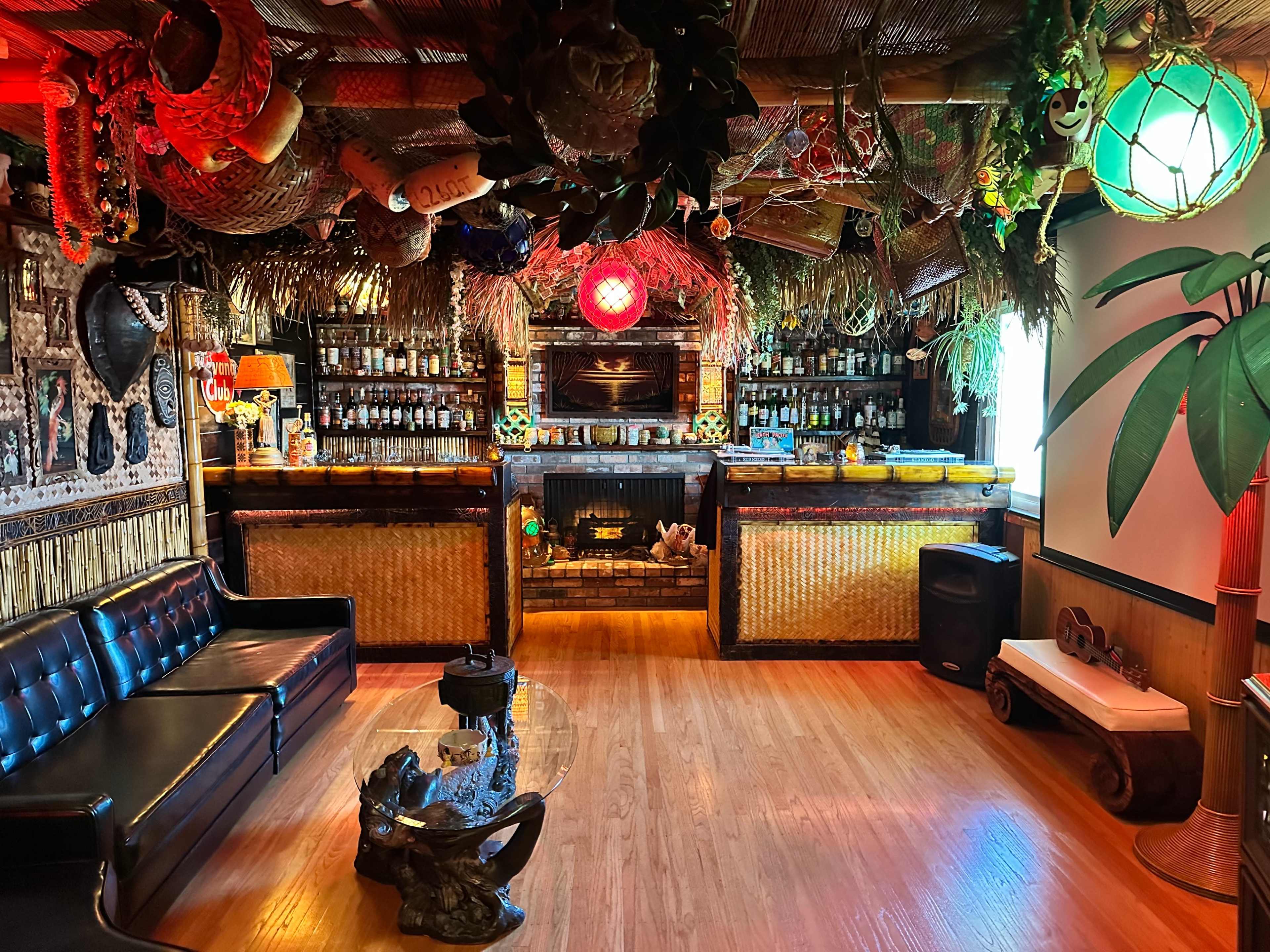 The image shows a tropical-themed bar with thatched decor, a wooden counter filled with bottles, and cozy seating arranged around a glass table.