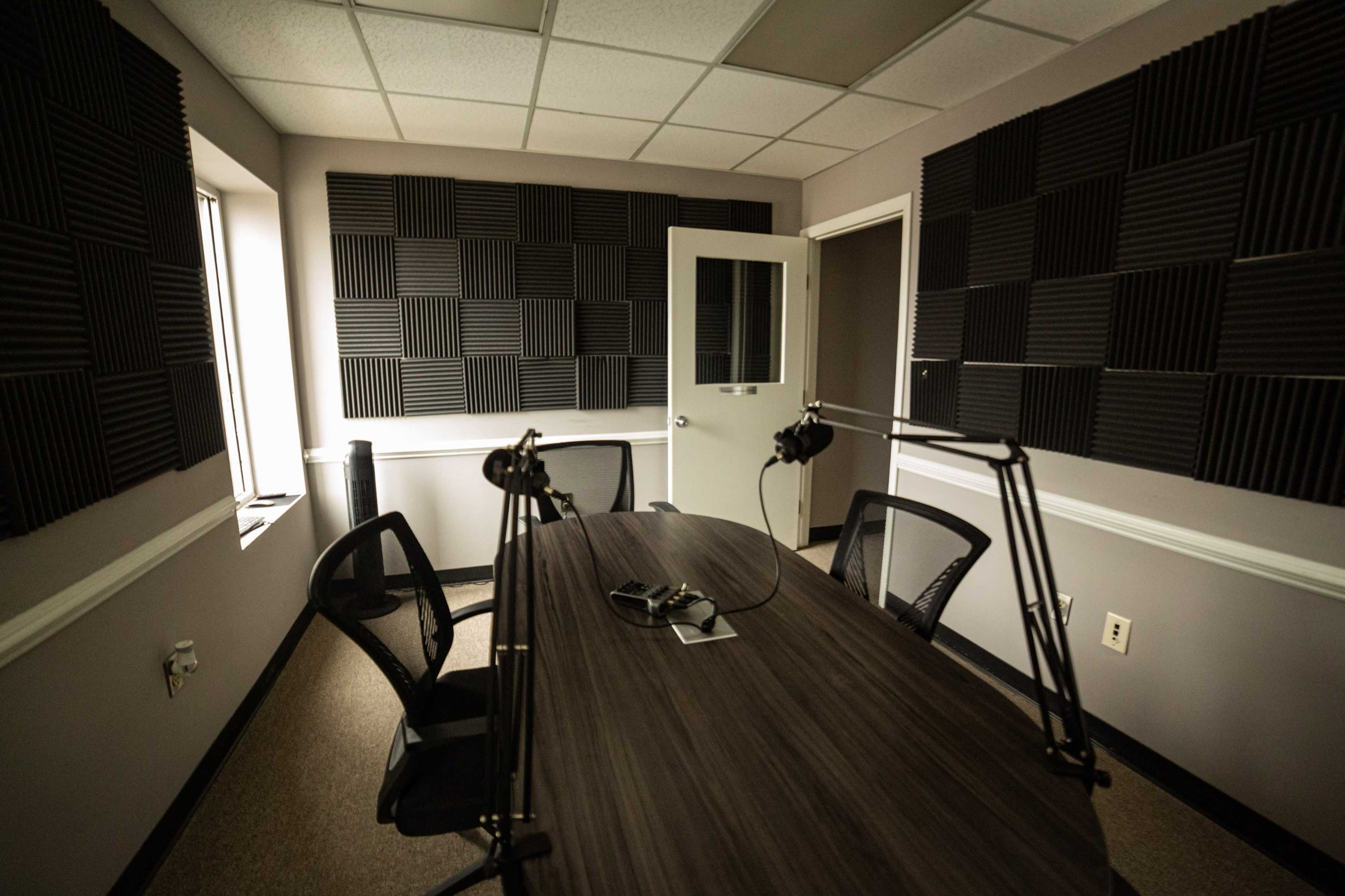 The image shows a recording studio with a large table, several chairs, and soundproof foam panels on the walls.