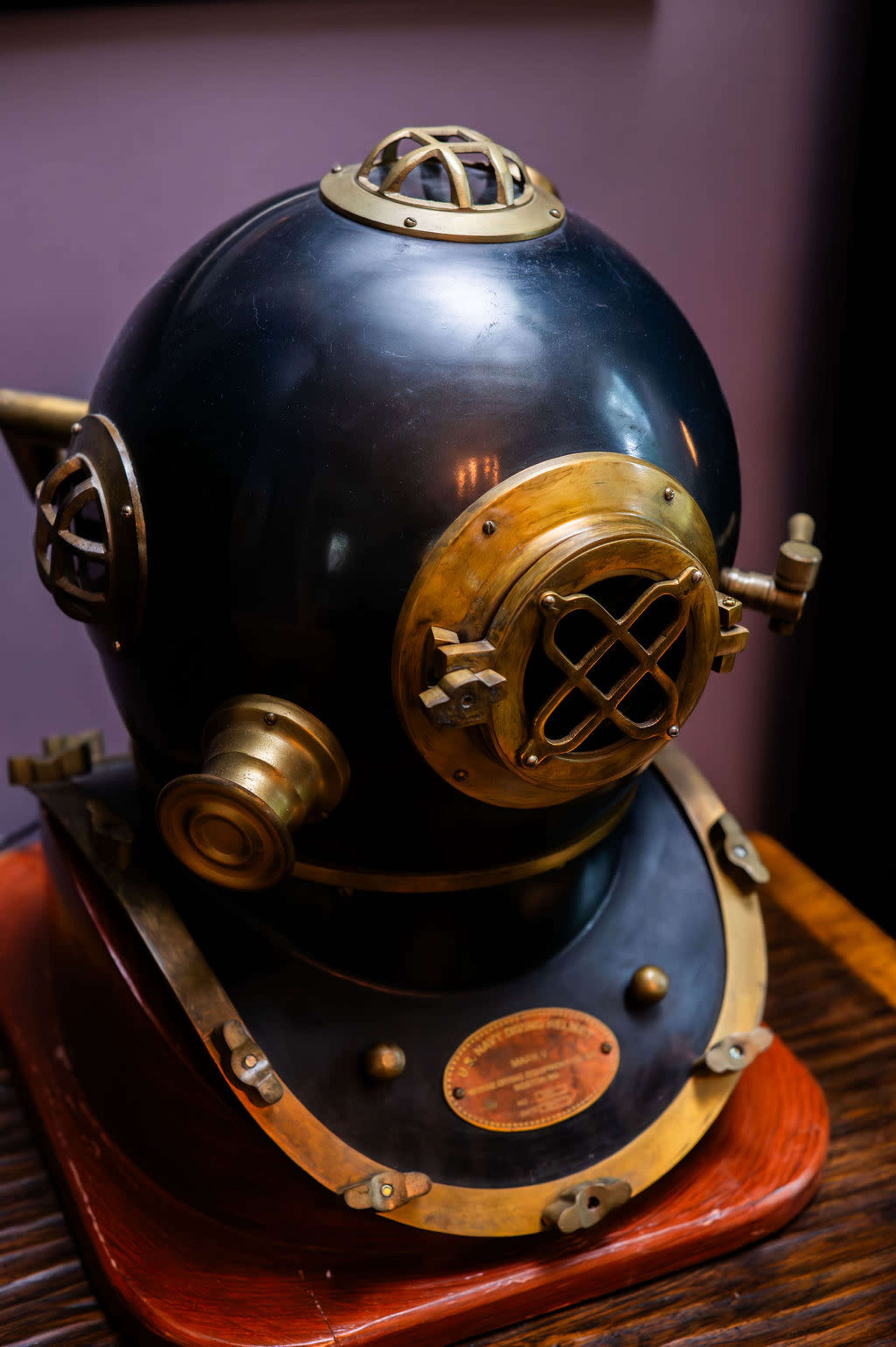 The image shows a vintage brass and black diving helmet resting on a wooden base.
