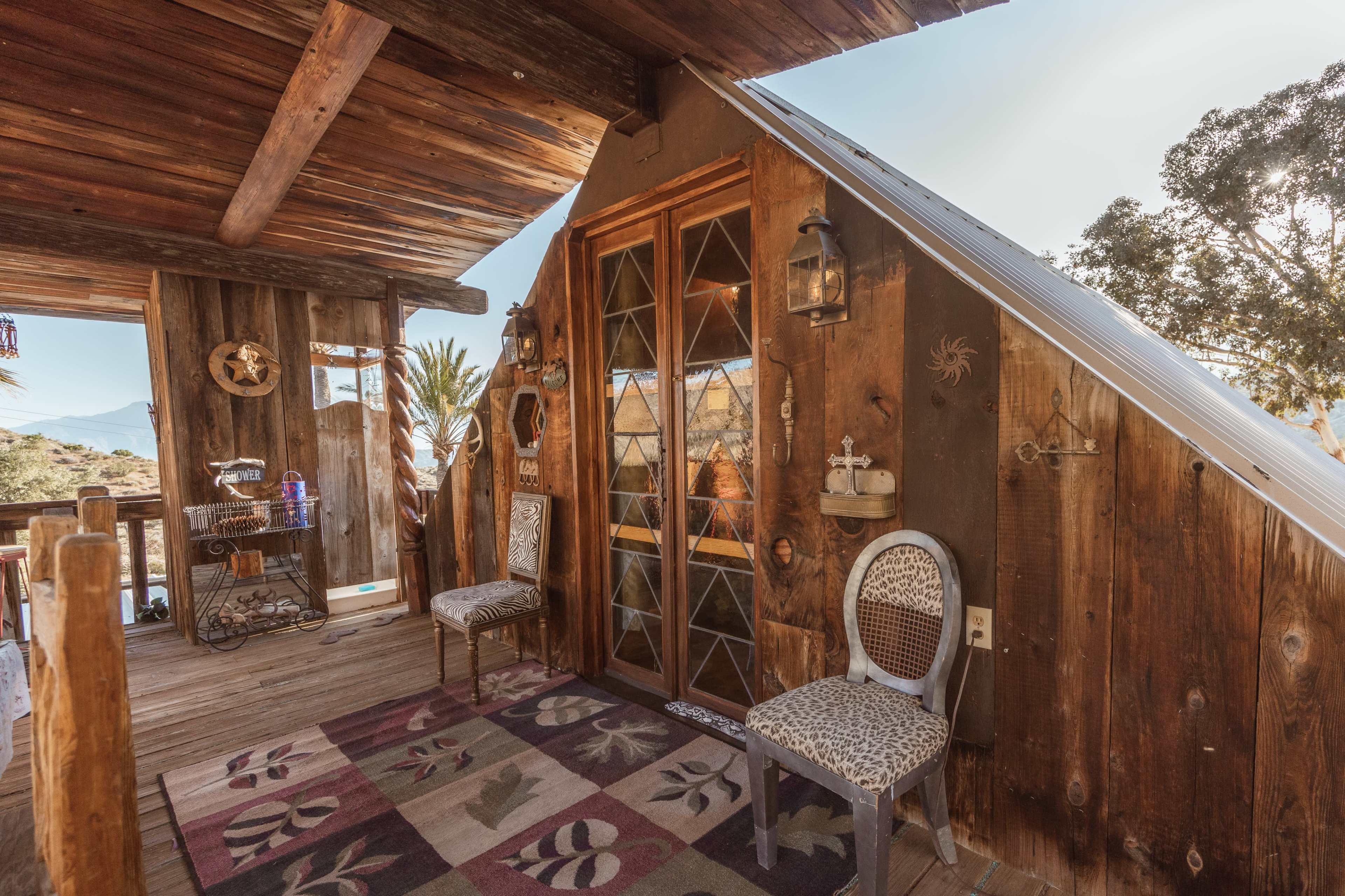 The image shows a rustic wooden balcony with two chairs and a door leading into a cabin or house.