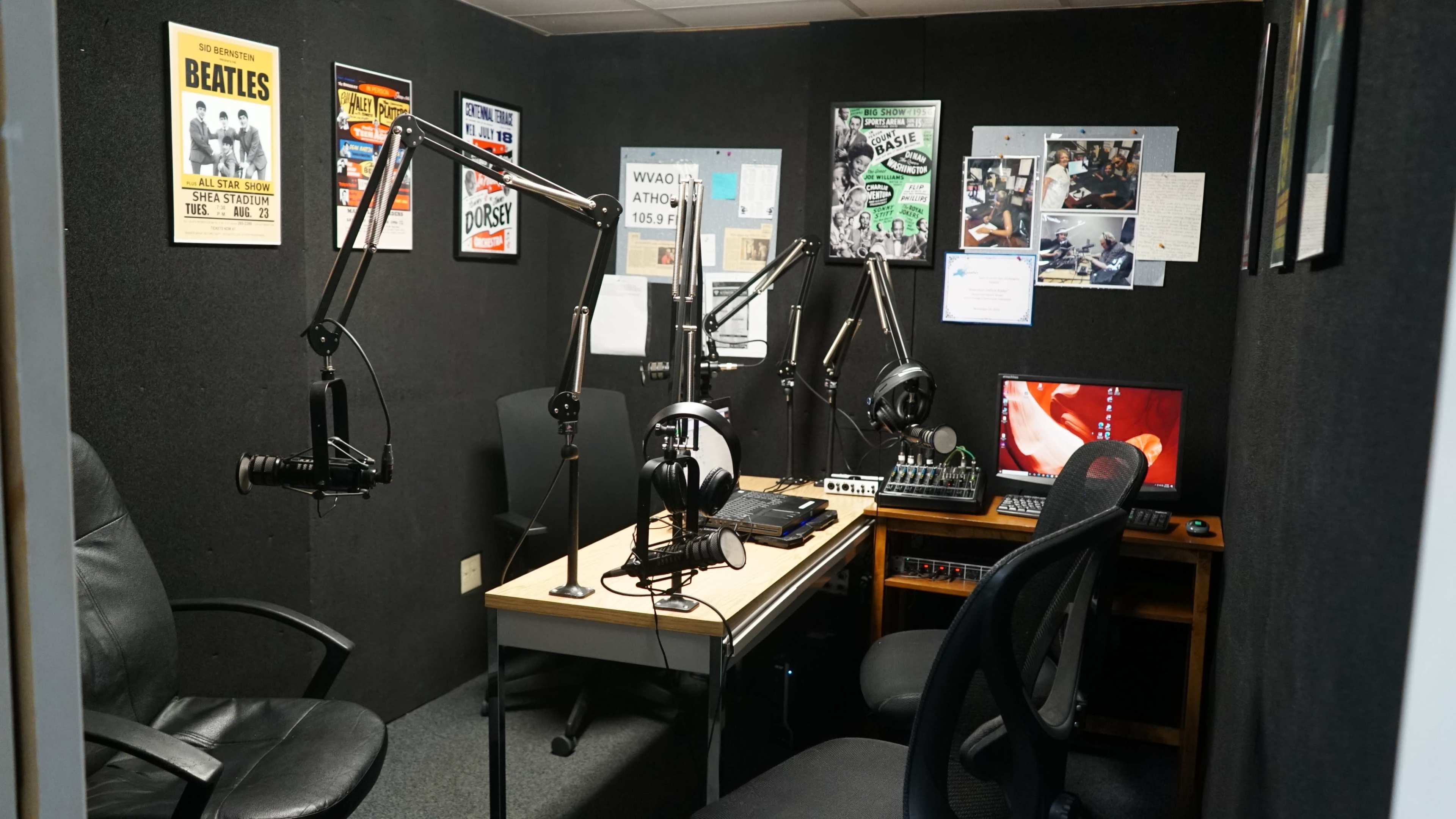 The image shows a small radio studio with two microphone stands, headphones, a sound mixer, and various posters on the walls.