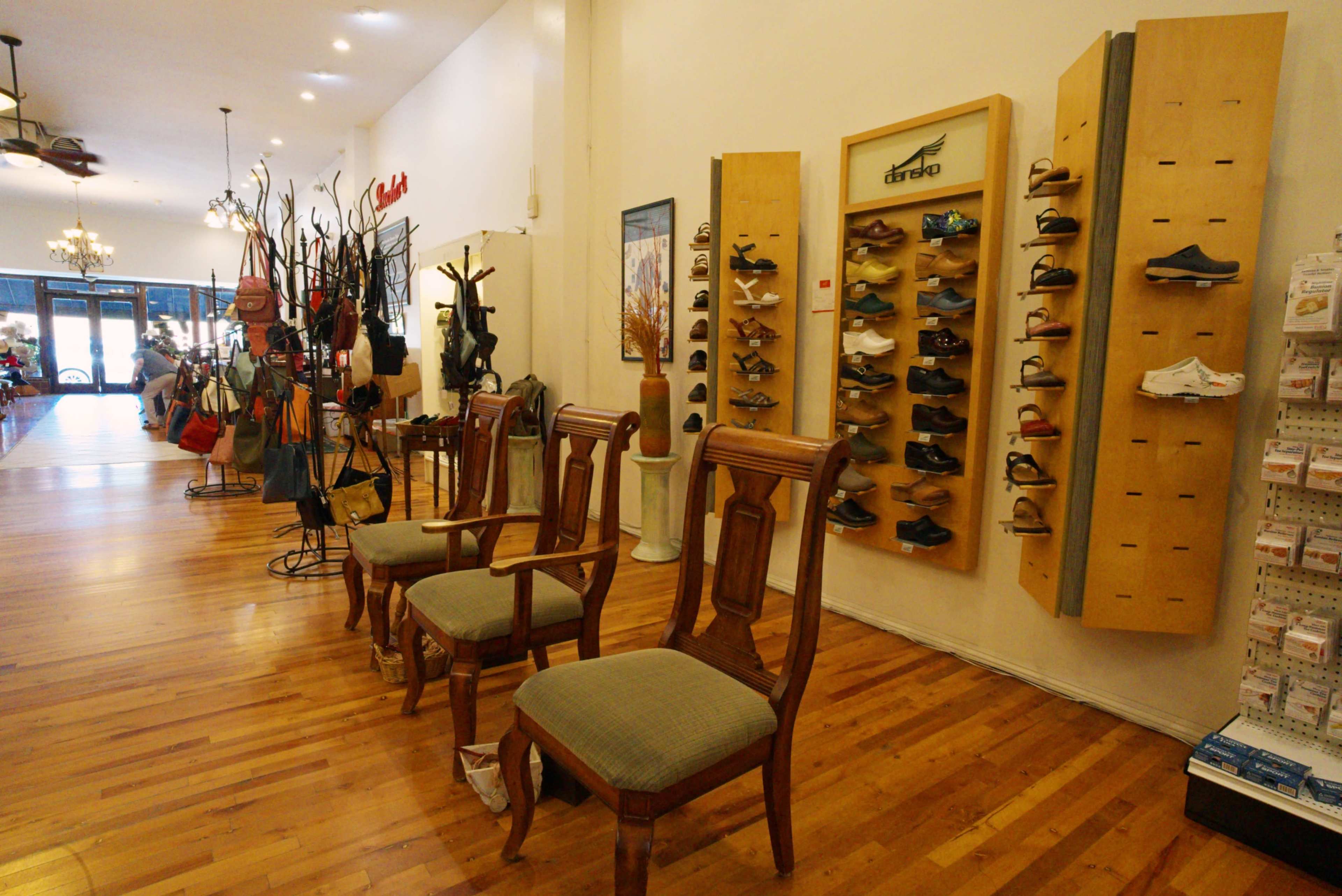 The image shows a retail space with wooden flooring, featuring display shelves for shoes on the wall, wooden chairs arranged in a row, and various bags and accessories hanging on the left side.