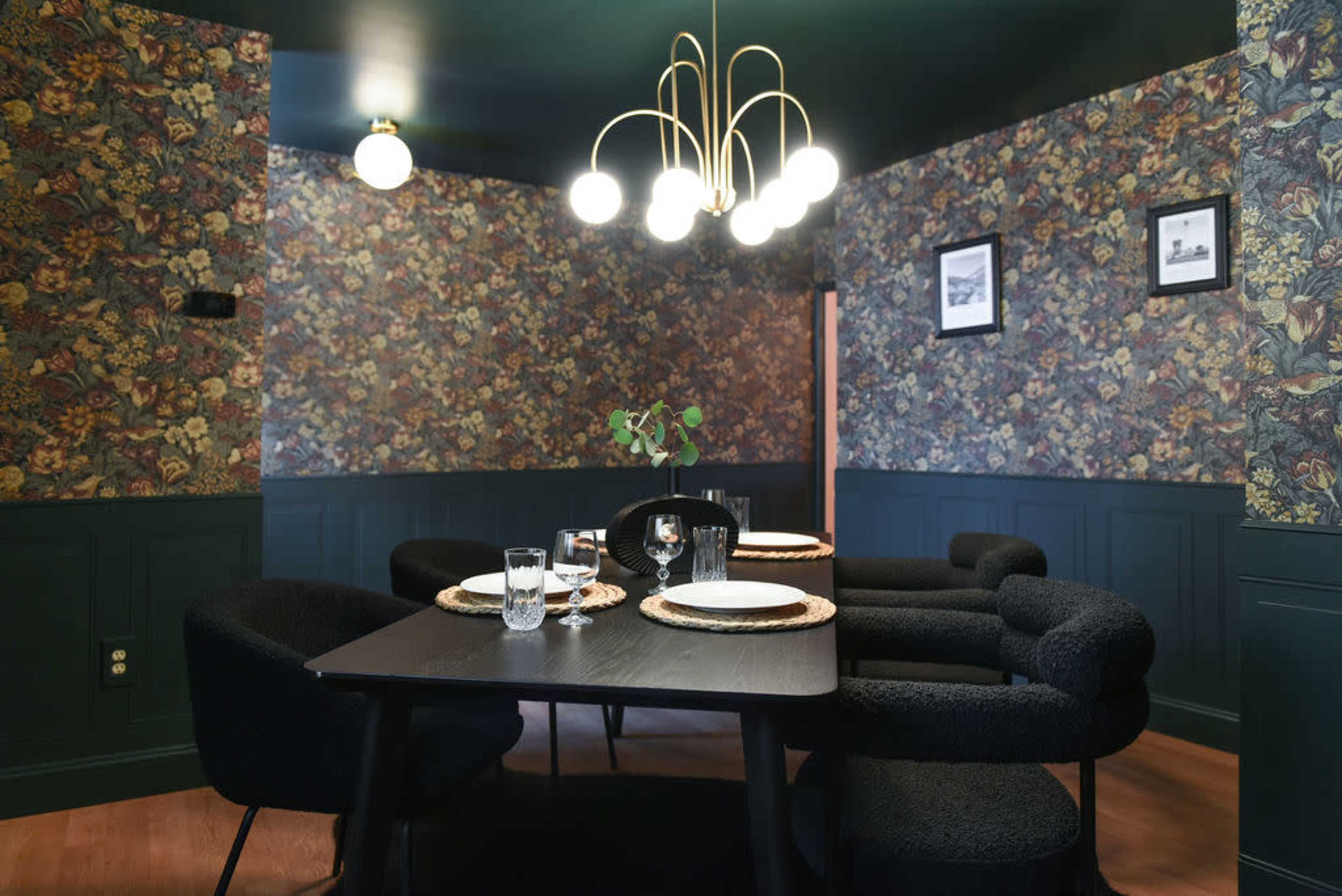 The image shows a dining area with a dark floral wallpaper, a black table surrounded by four upholstered chairs, and a modern light fixture hanging from the ceiling.