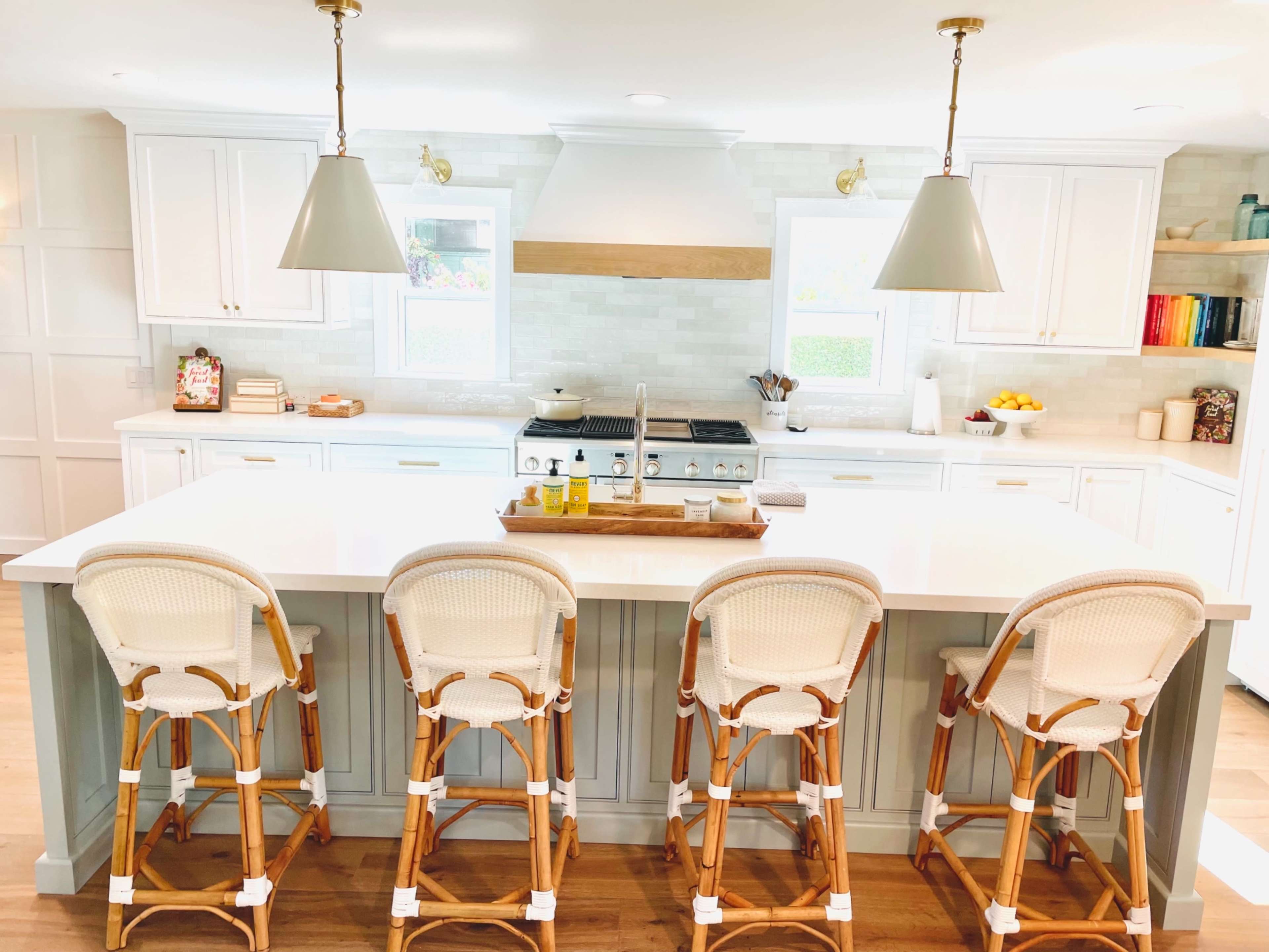 A modern kitchen with a large island featuring four wicker barstools, white cabinetry, a gas stove, and pendant lighting.