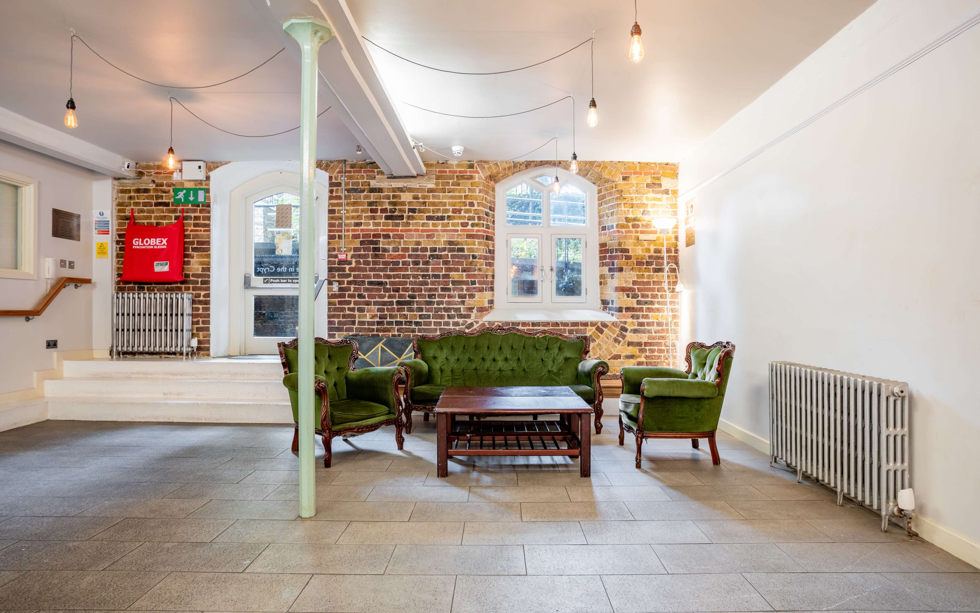 The interior features a cozy seating area with a green velvet sofa and chairs arranged around a wooden coffee table, accented by a brick wall and large windows.