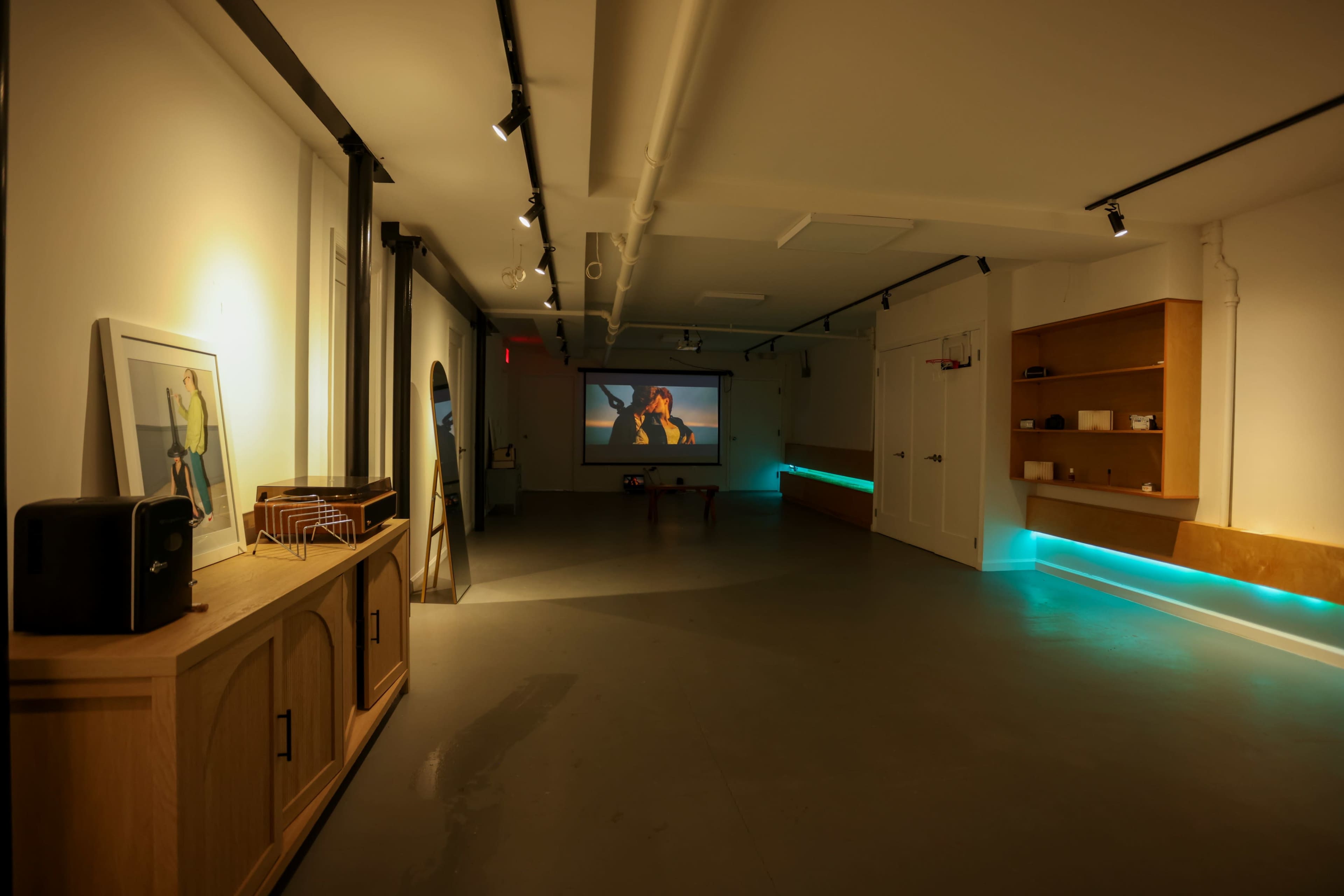 A spacious, well-lit room features a large projector screen displaying a film, with modern furniture and blue LED lighting along the walls.