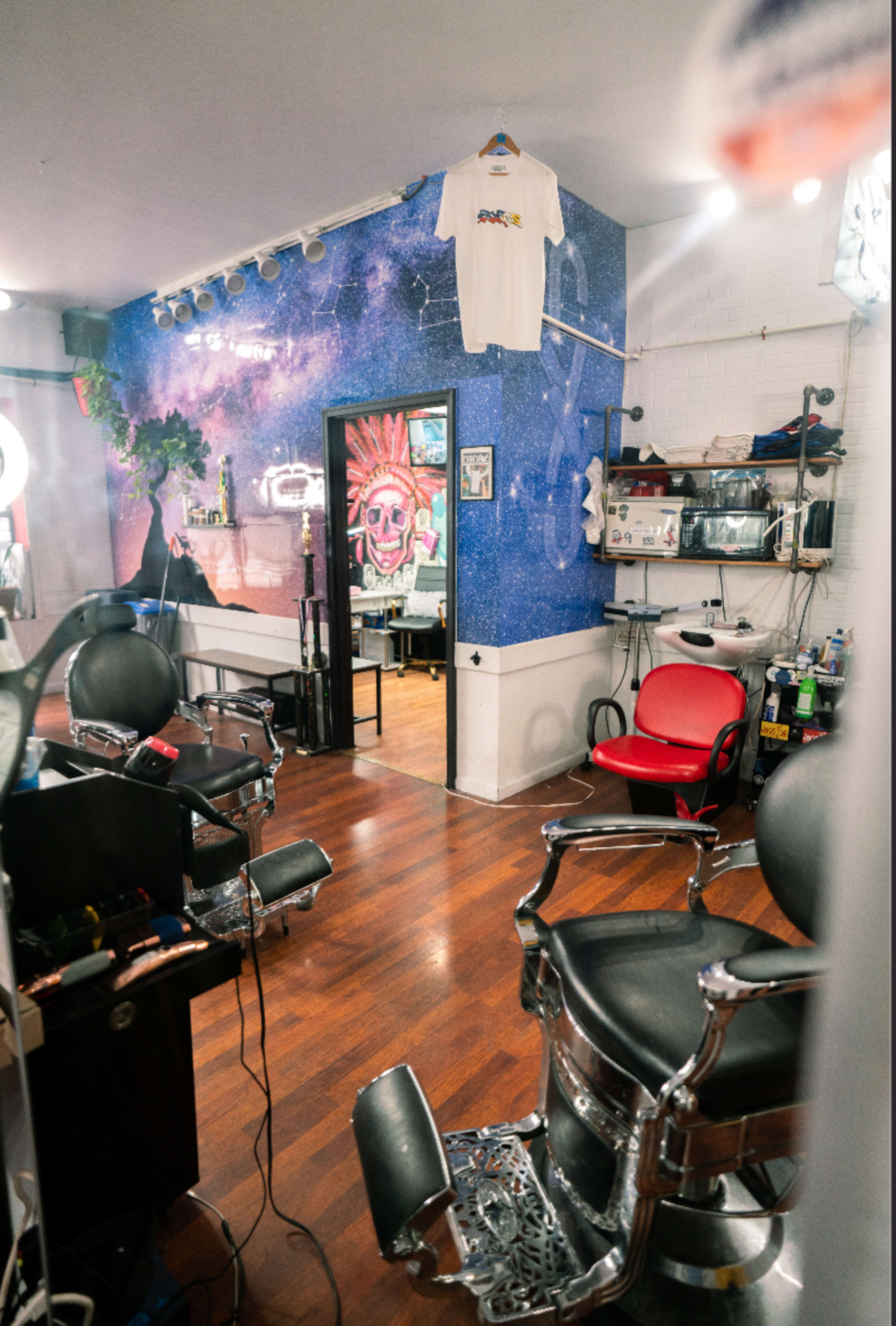 The image shows a brightly decorated barbershop with black barber chairs, a red chair, and a mural featuring a cosmic scene on one wall.