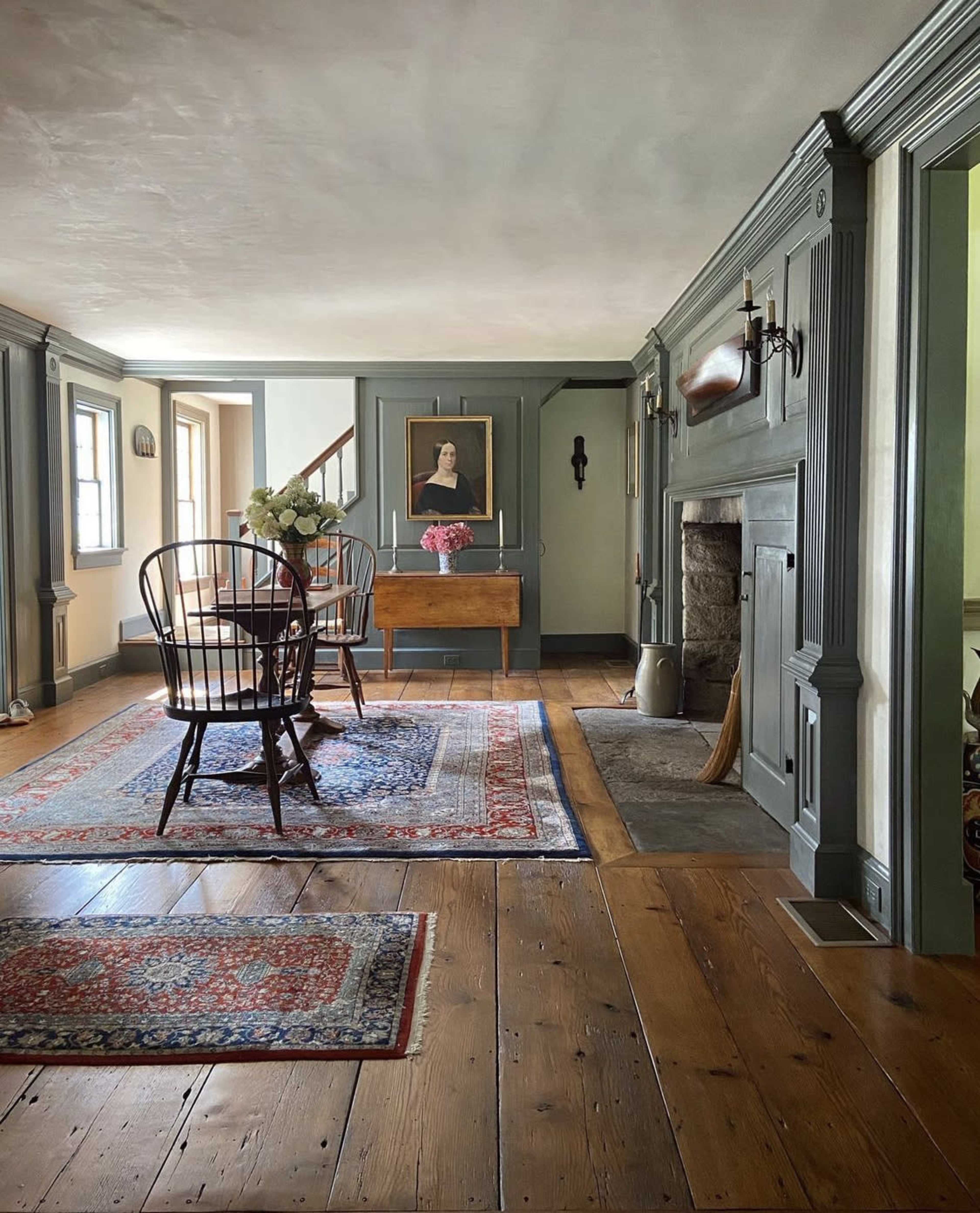 A room with wooden flooring, a fireplace, a table with a vase of flowers, and two black chairs arranged near a rug, with a portrait hanging on the wall.