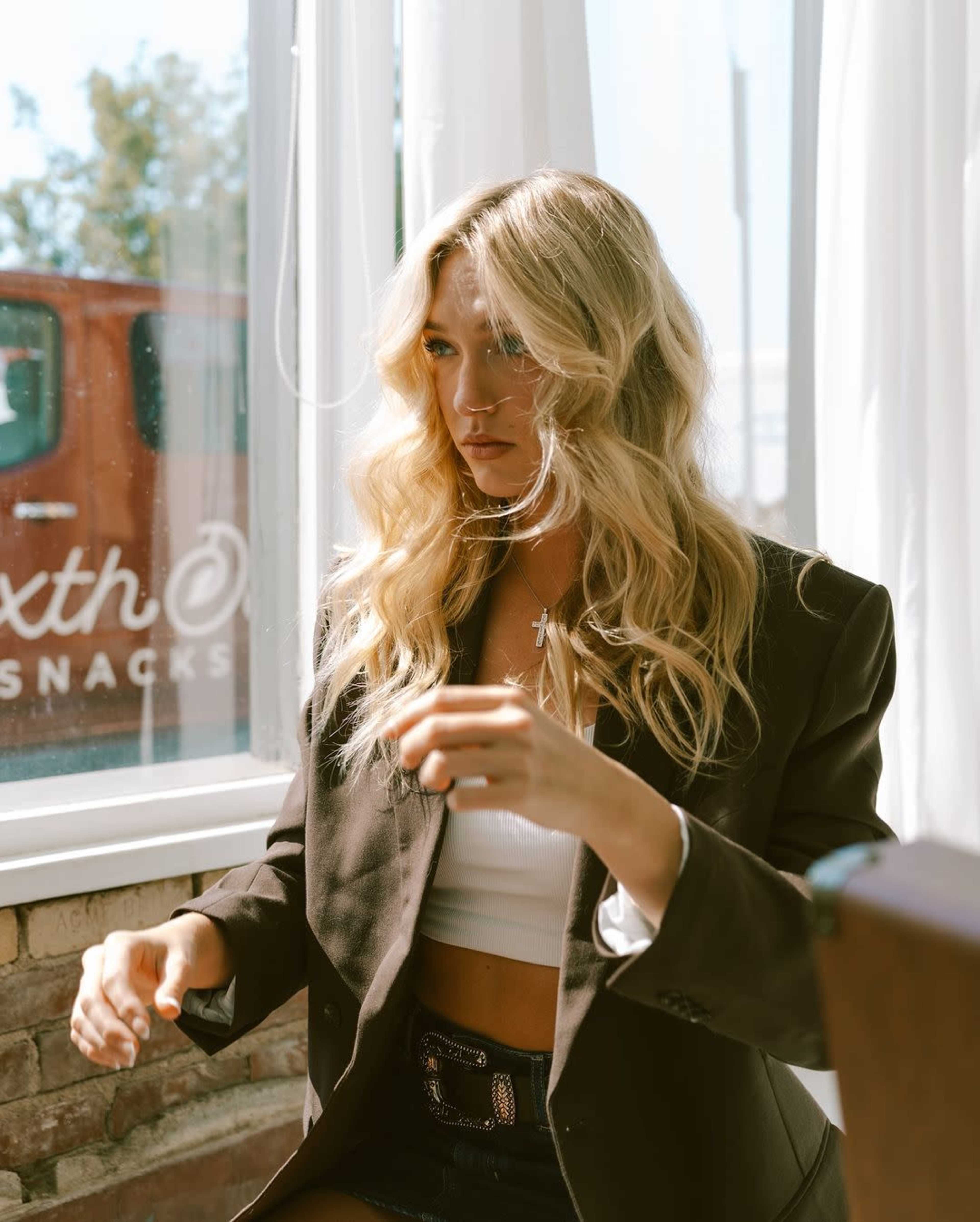 A person with long, wavy hair sits at a table in a bright room, wearing a black blazer and a white crop top, while looking thoughtfully out a window.