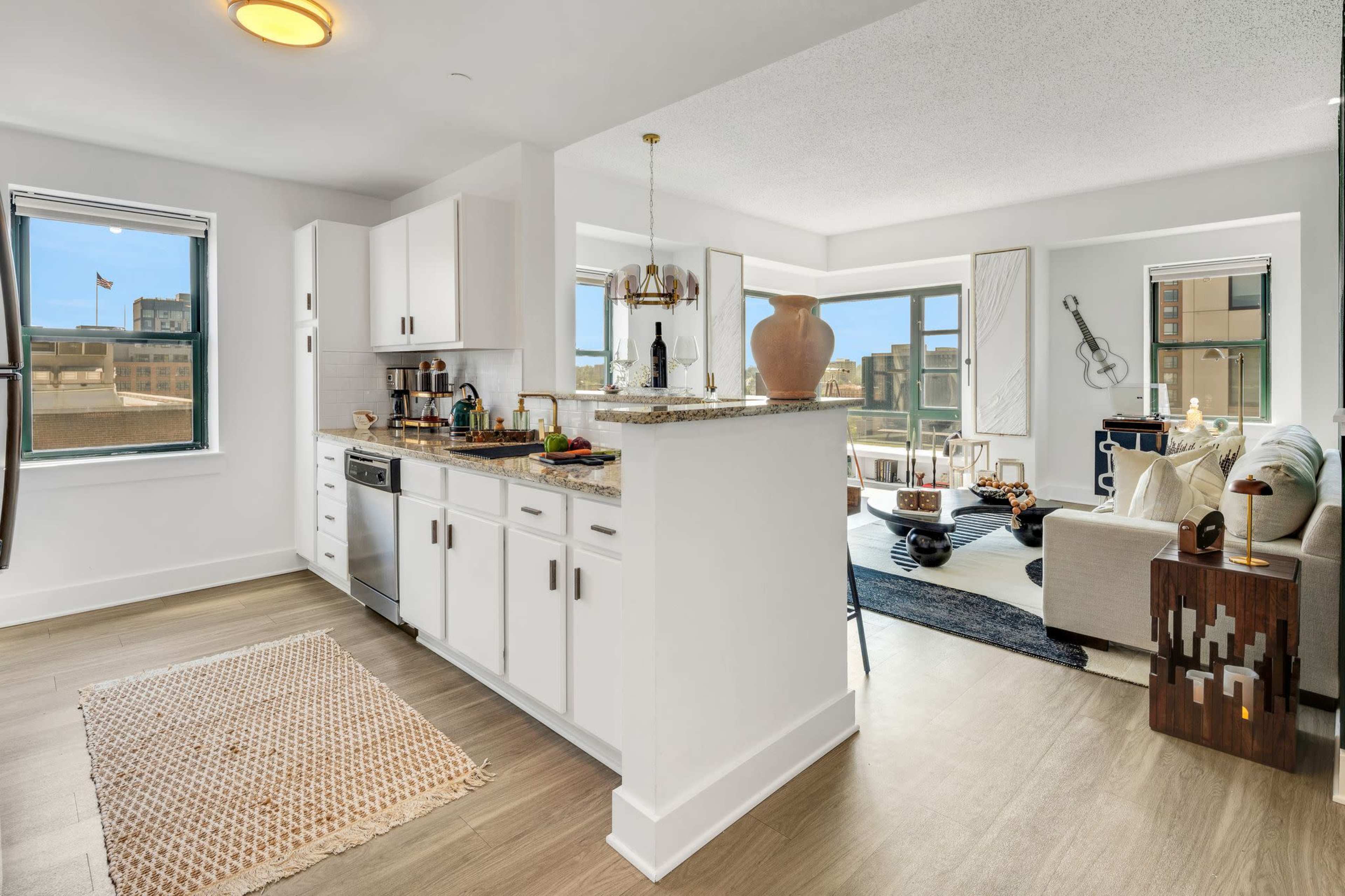 A modern open-concept kitchen and living area features white cabinets and a granite countertop, with large windows allowing natural light to fill the space.