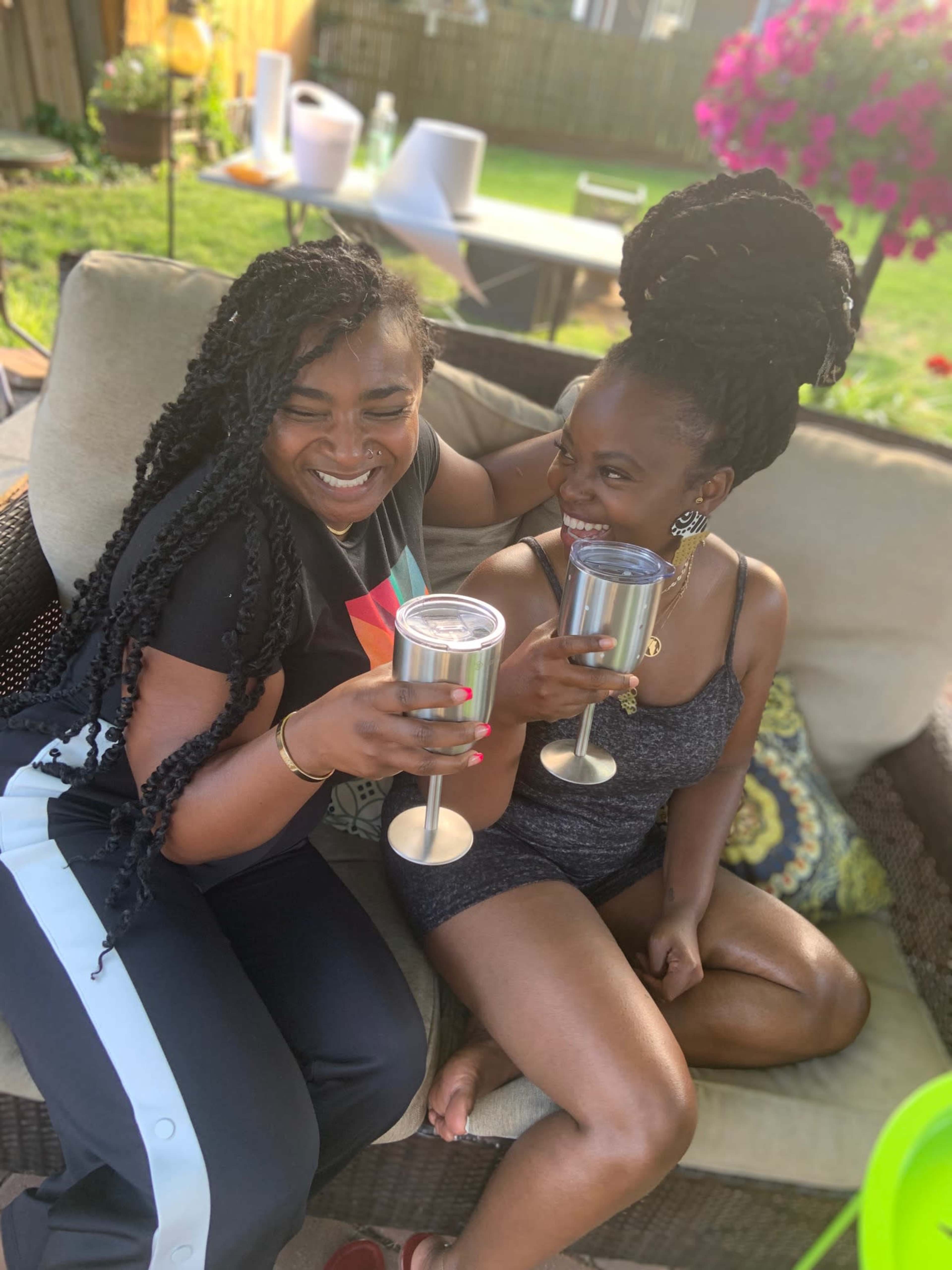 Two women are sitting on a patio couch, clinking stainless steel cups in celebration.
