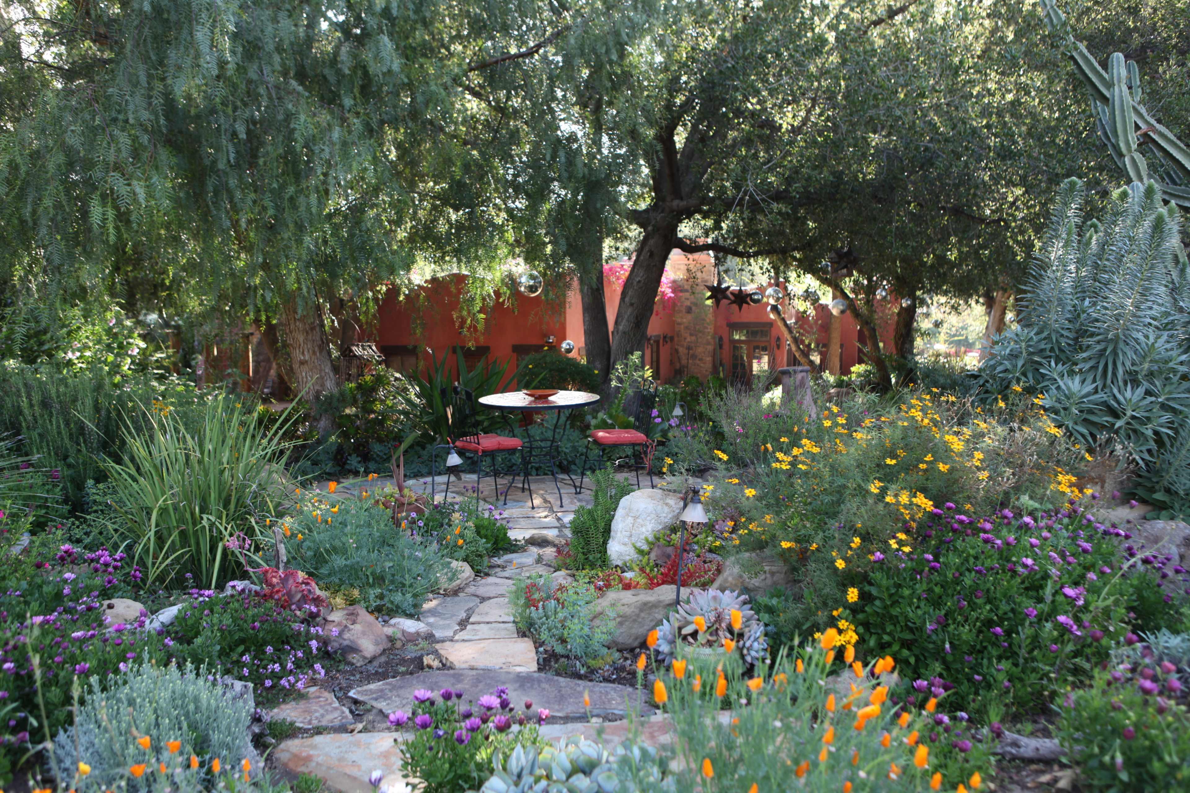 A stone pathway leads through a vibrant garden filled with various colorful flowers and greenery, with a small table and chairs set under the trees.