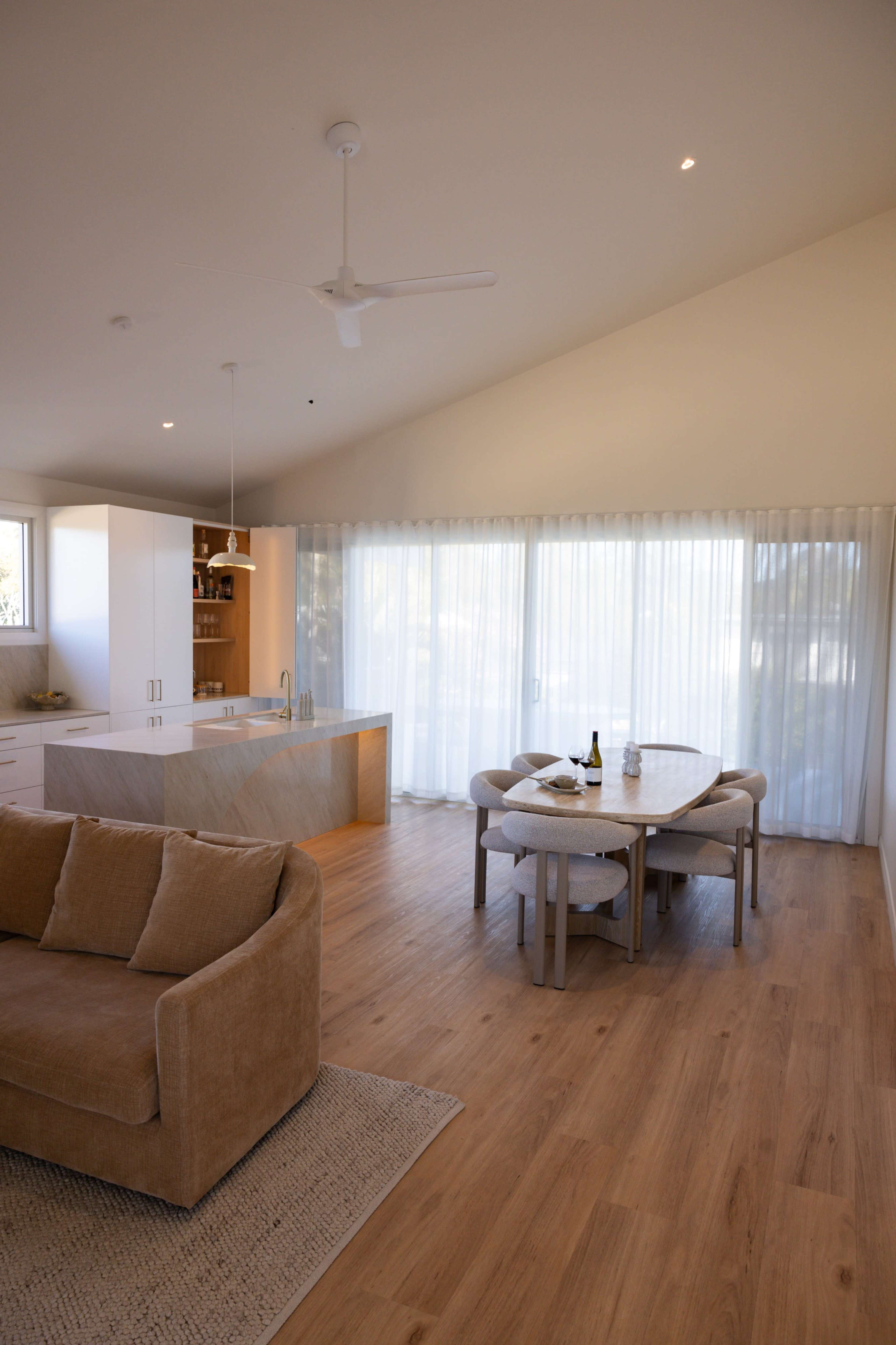 A spacious, modern interior features a kitchen island and a dining table surrounded by chairs, with large windows covered by sheer curtains allowing natural light to enter.