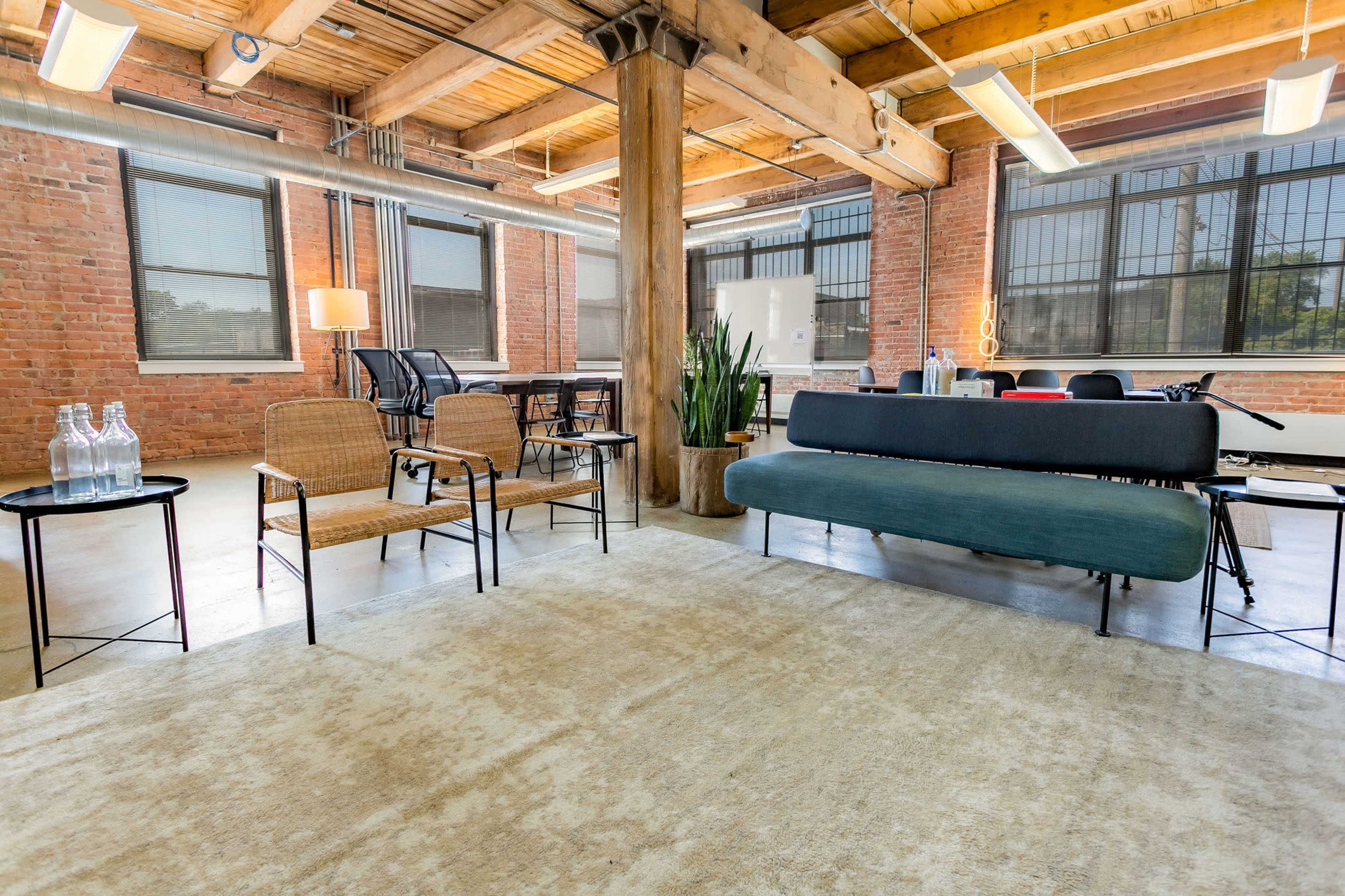 The image shows a spacious industrial-style office with exposed brick walls, wooden beams, and various seating arrangements, including a sofa and chairs, along with a rug and decorative plants.