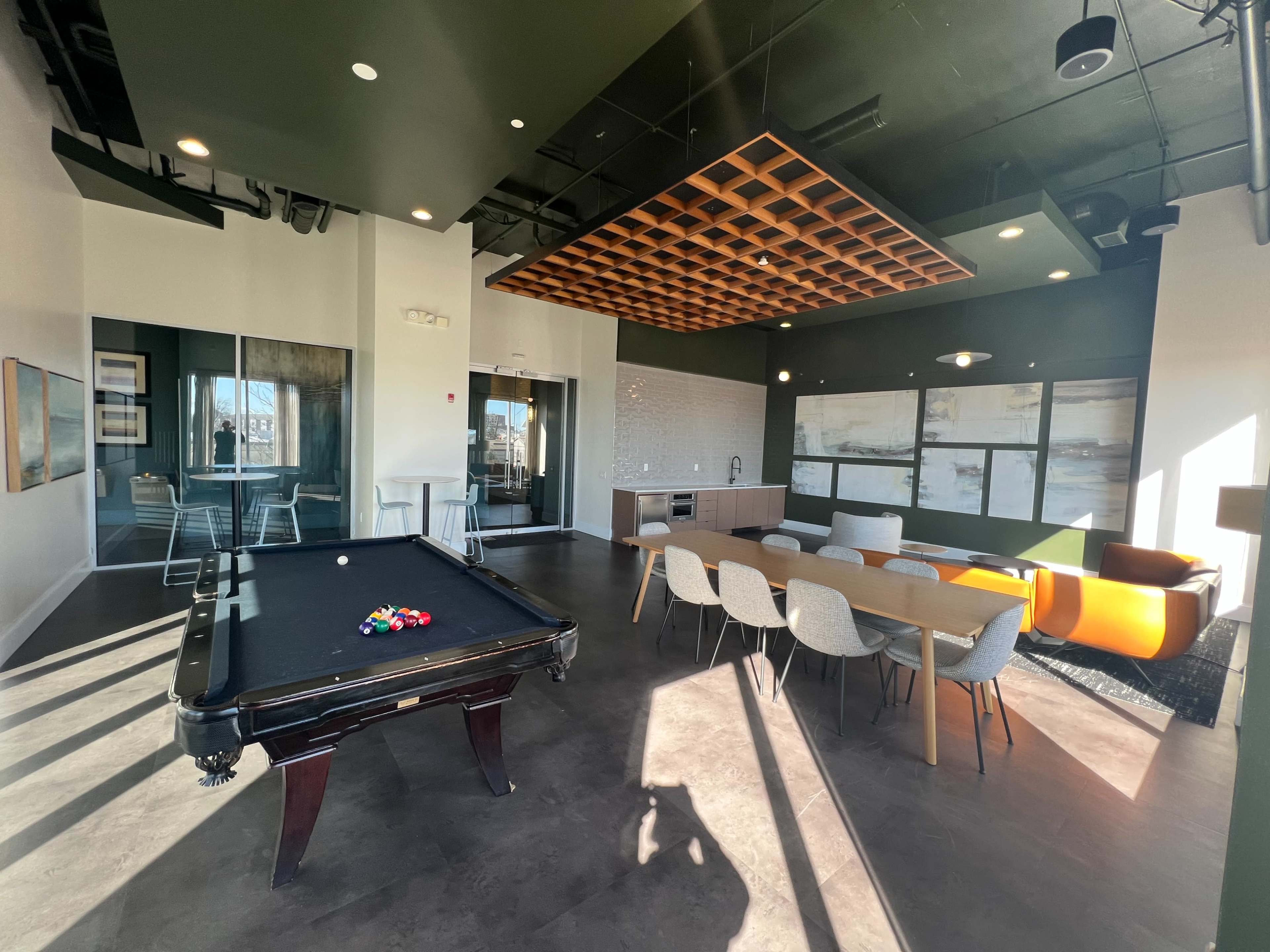 A modern lounge area featuring a pool table, a dining table, and seating arrangements with large windows allowing natural light.
