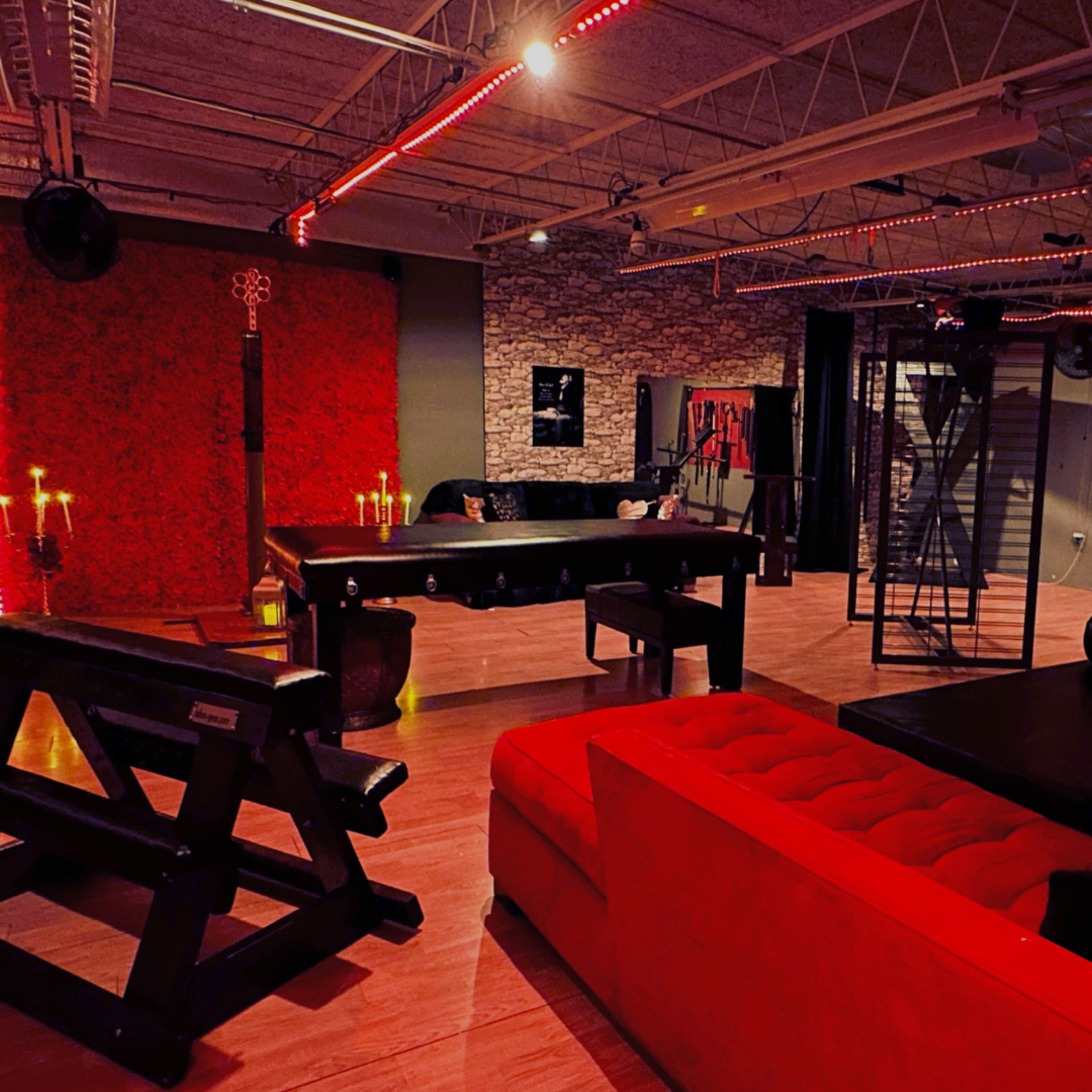 The image shows a dimly lit room featuring a red wall, a black couch, and a large table, with red lighting and various decorative elements.