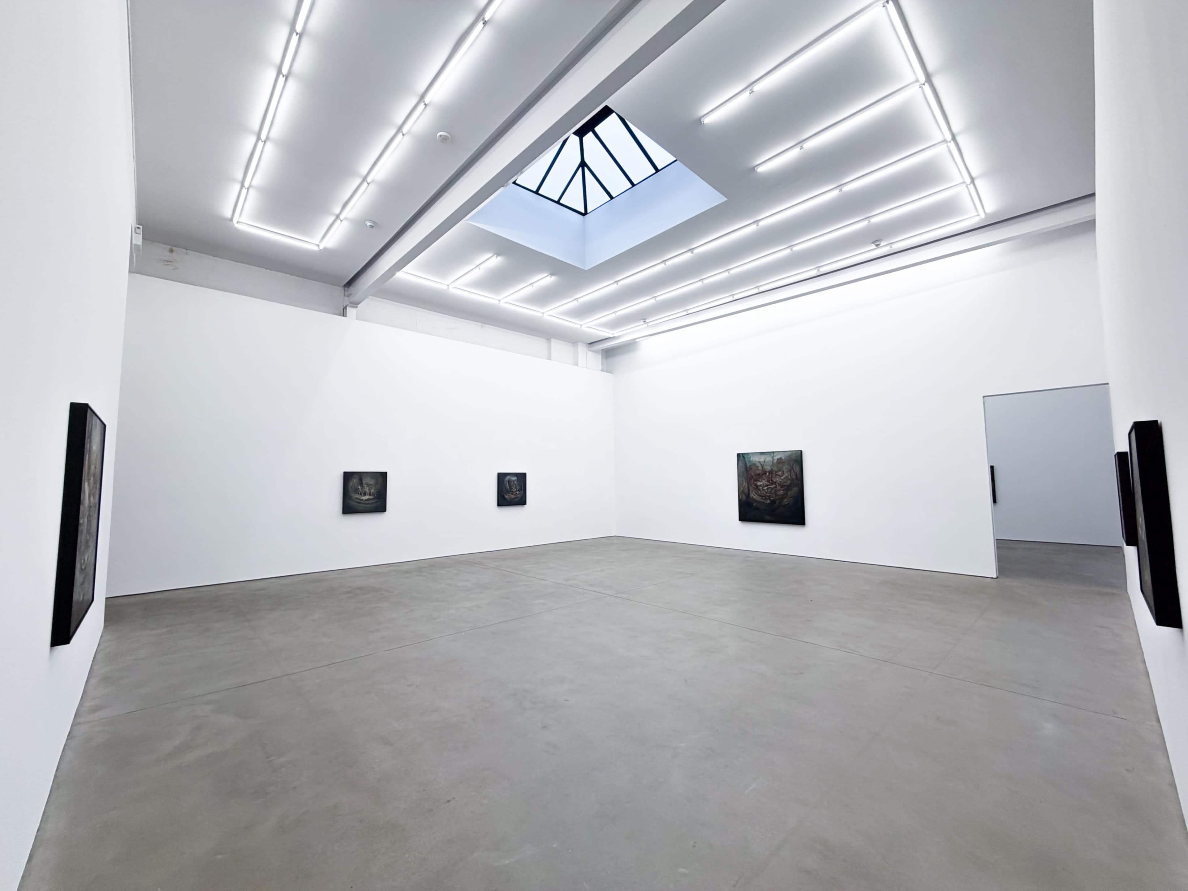 The image shows a spacious art gallery with white walls, featuring several framed artworks evenly spaced along the walls and a skylight above.