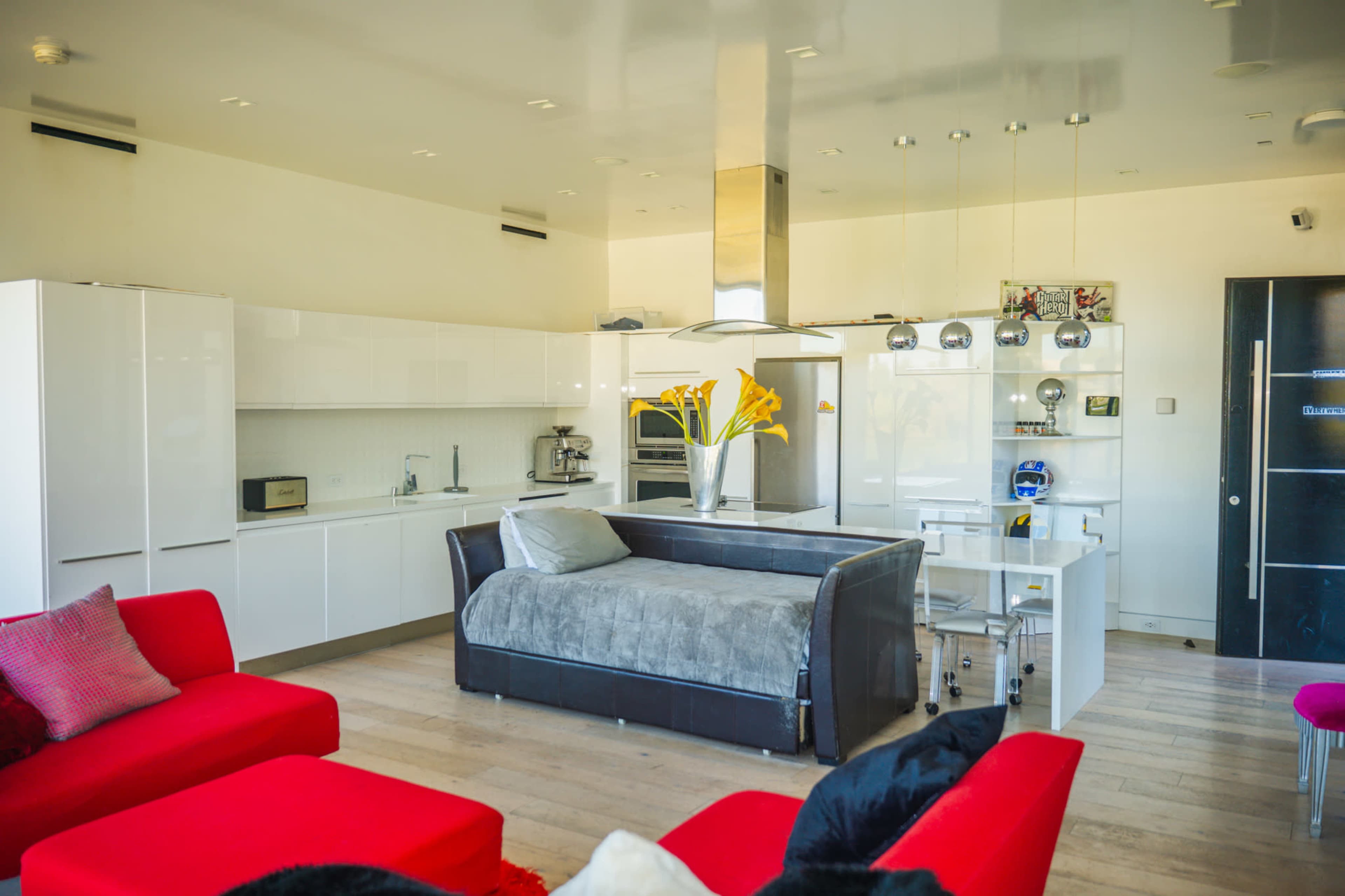 A modern kitchen and living area features sleek white cabinets, a central gray sofa, and bright red seating, with a stainless steel range hood and a glass dining table.