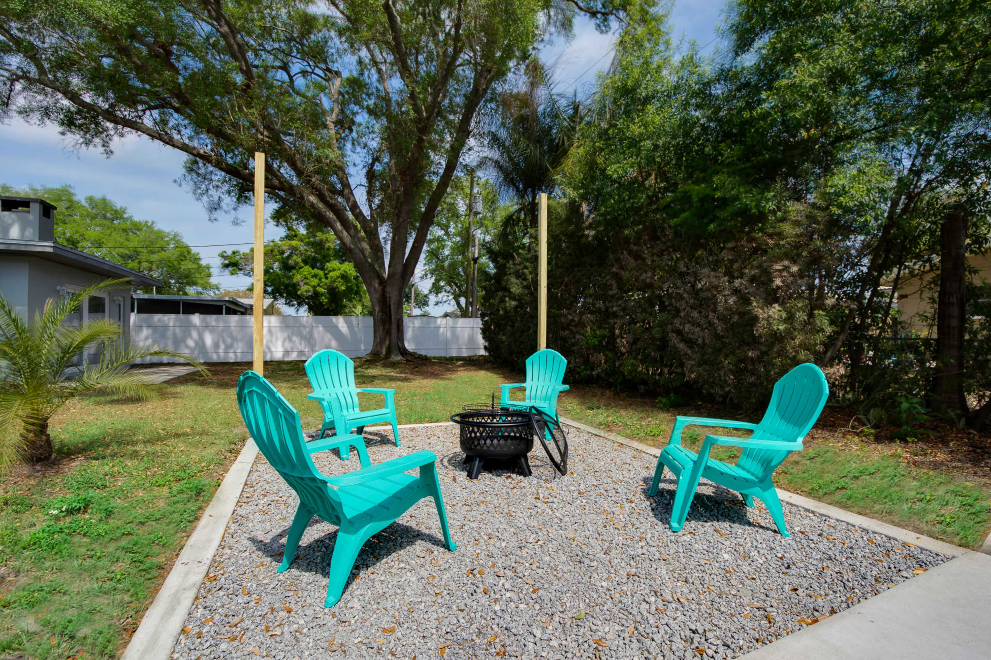 A backyard with four turquoise chairs arranged around a fire pit on a gravel area, surrounded by greenery and trees.