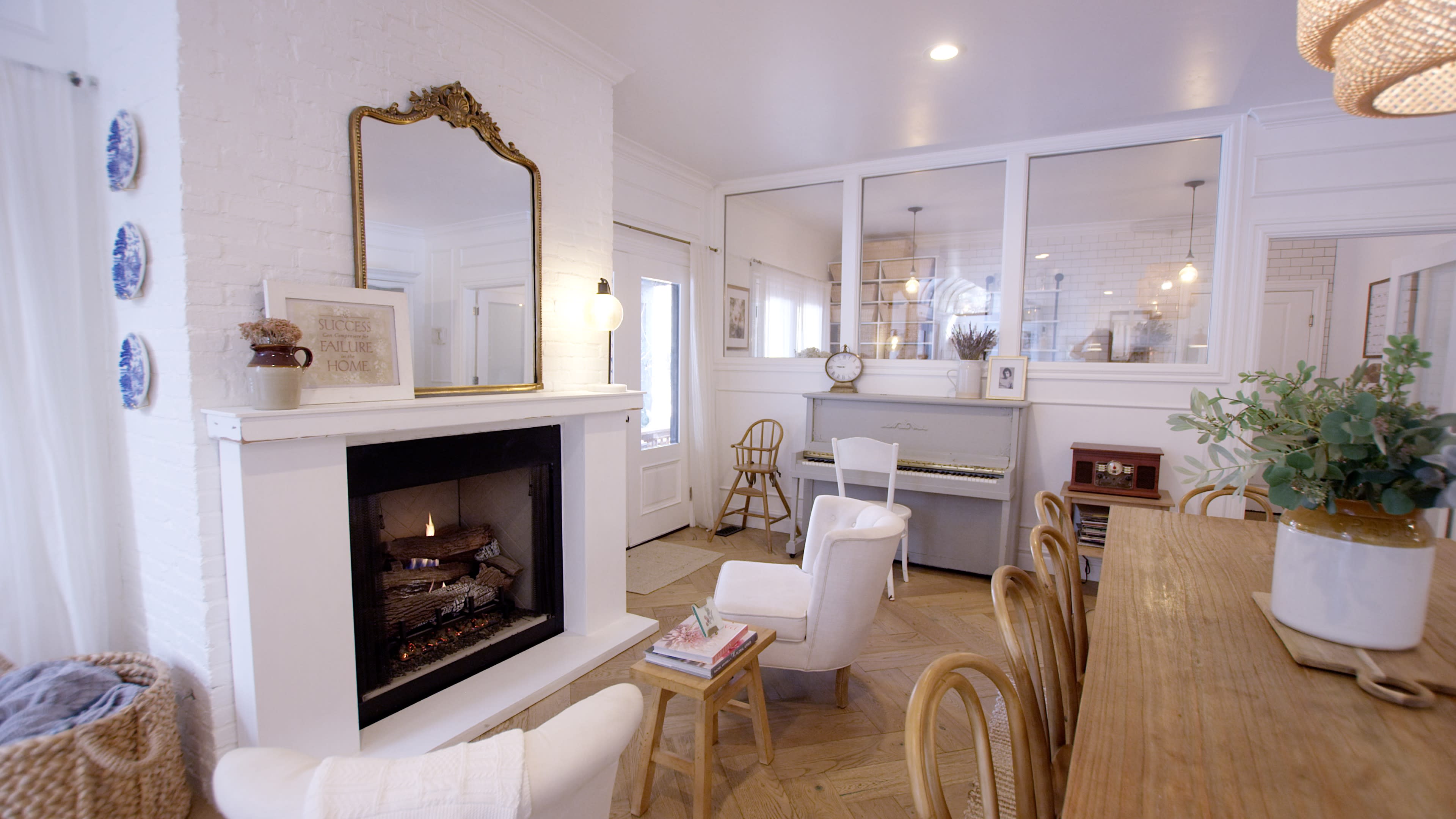 A cozy living space featuring a fireplace with a mirror above it, surrounded by white walls, a dining table, and a piano in the background.