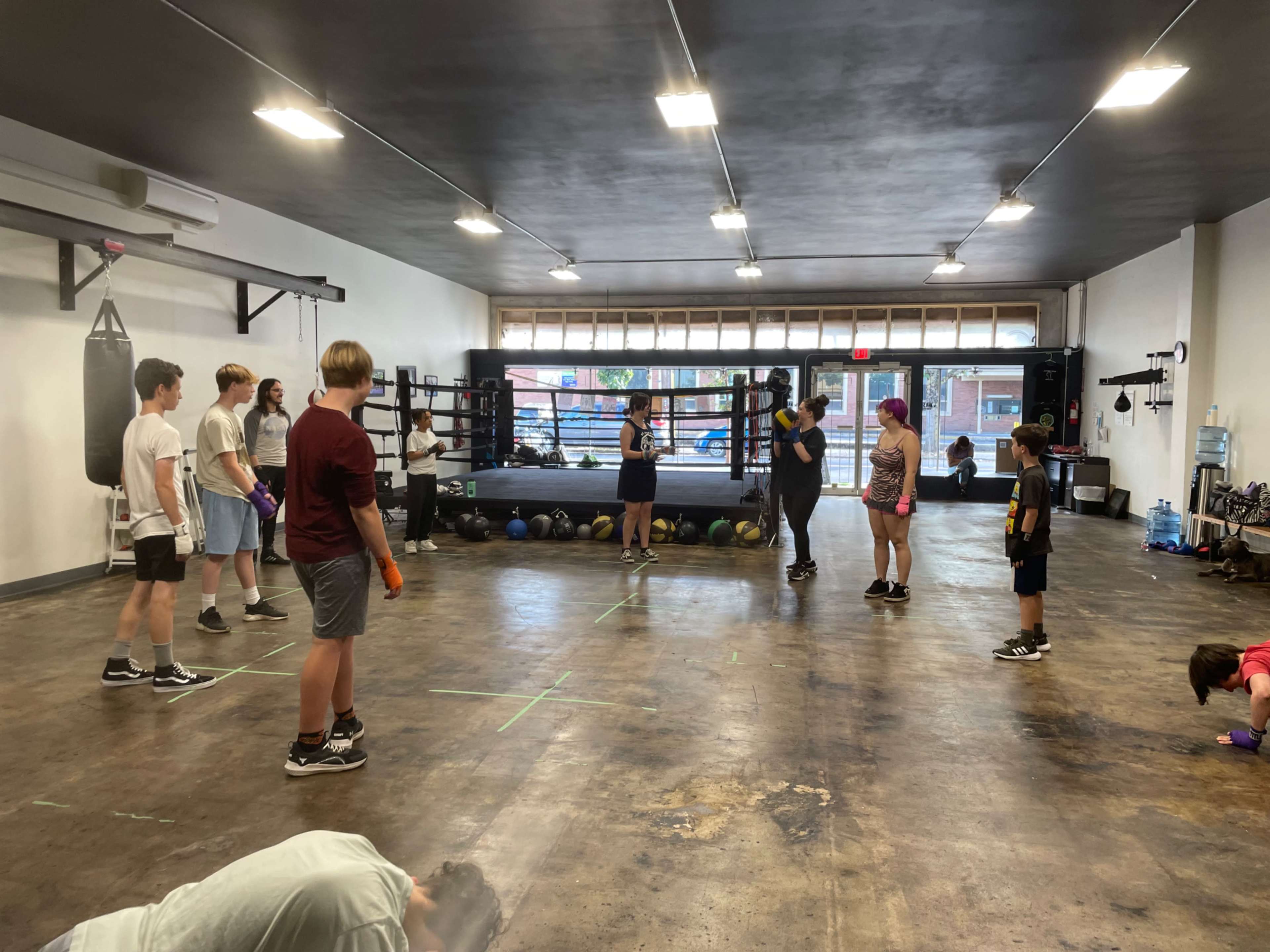 A group of young people participates in a boxing training session inside a spacious gym featuring a boxing ring and various training equipment.