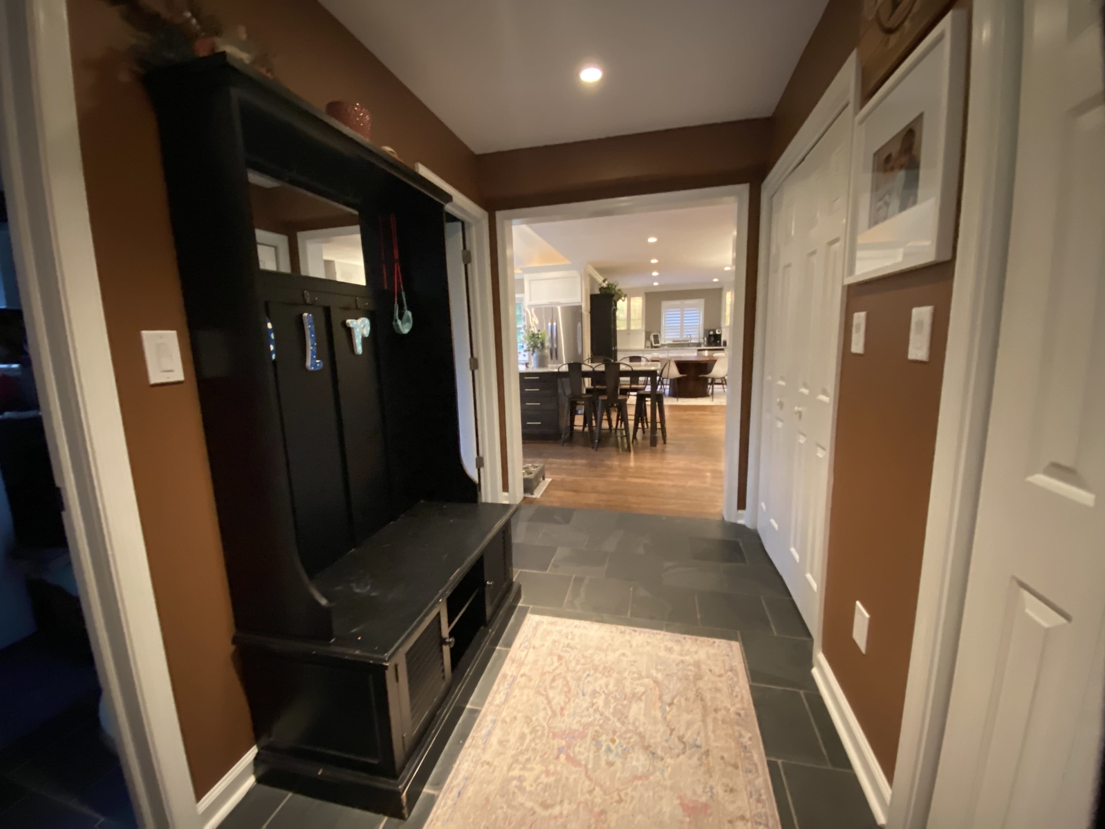 A hallway with a dark entryway bench, slate flooring, and a view into a warm-colored kitchen area beyond.