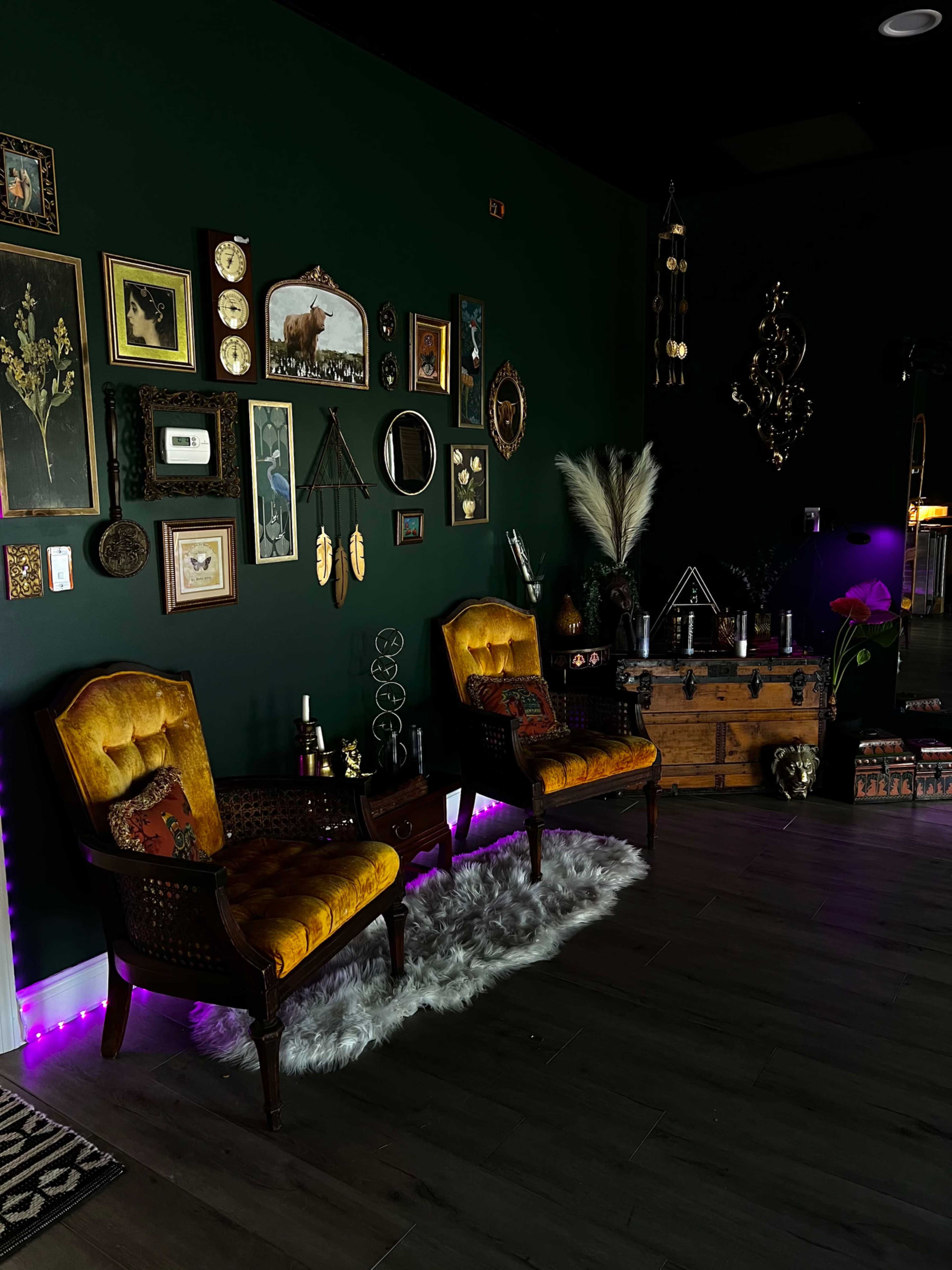 A dimly lit room featuring two vintage yellow chairs beside a wooden trunk, adorned with various framed artworks and decorative items on a dark green wall.