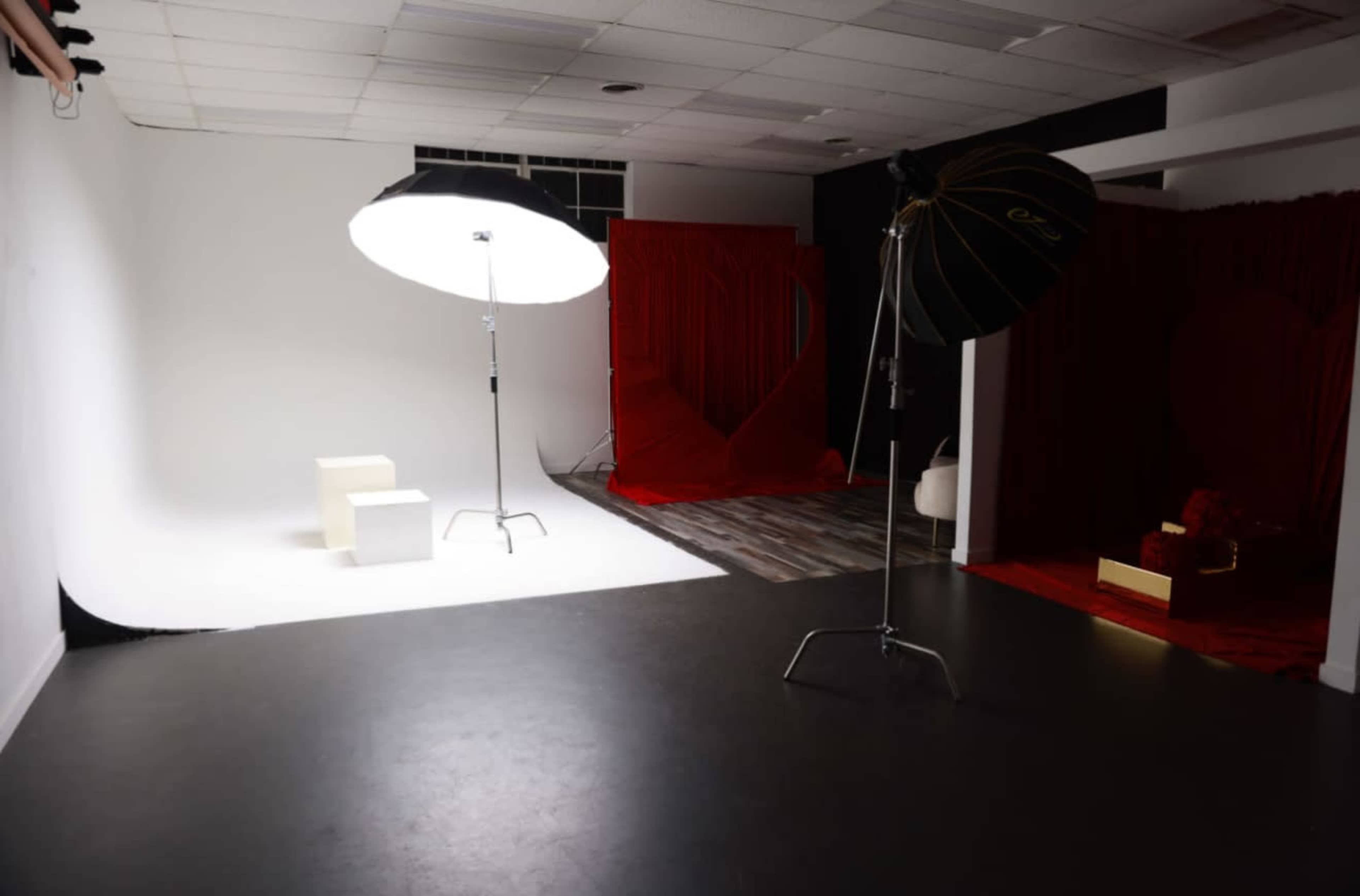The image shows a photography studio with a white backdrop, a large softbox light on a stand, and a red-themed setup in the background.
