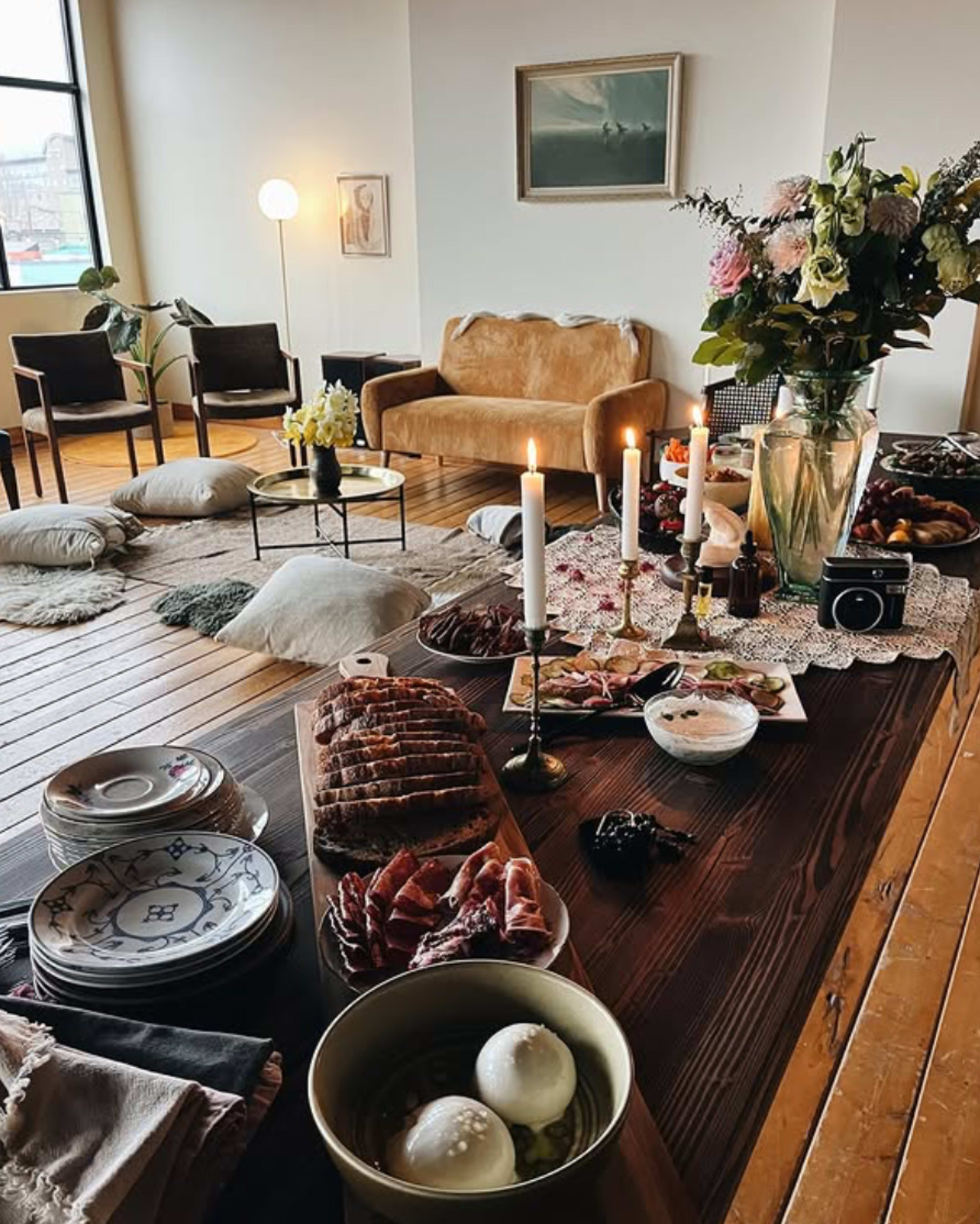 The scene features a cozy living room with a wooden dining table laid out with an array of food, candles, and decorative items, surrounded by seating and a large window.