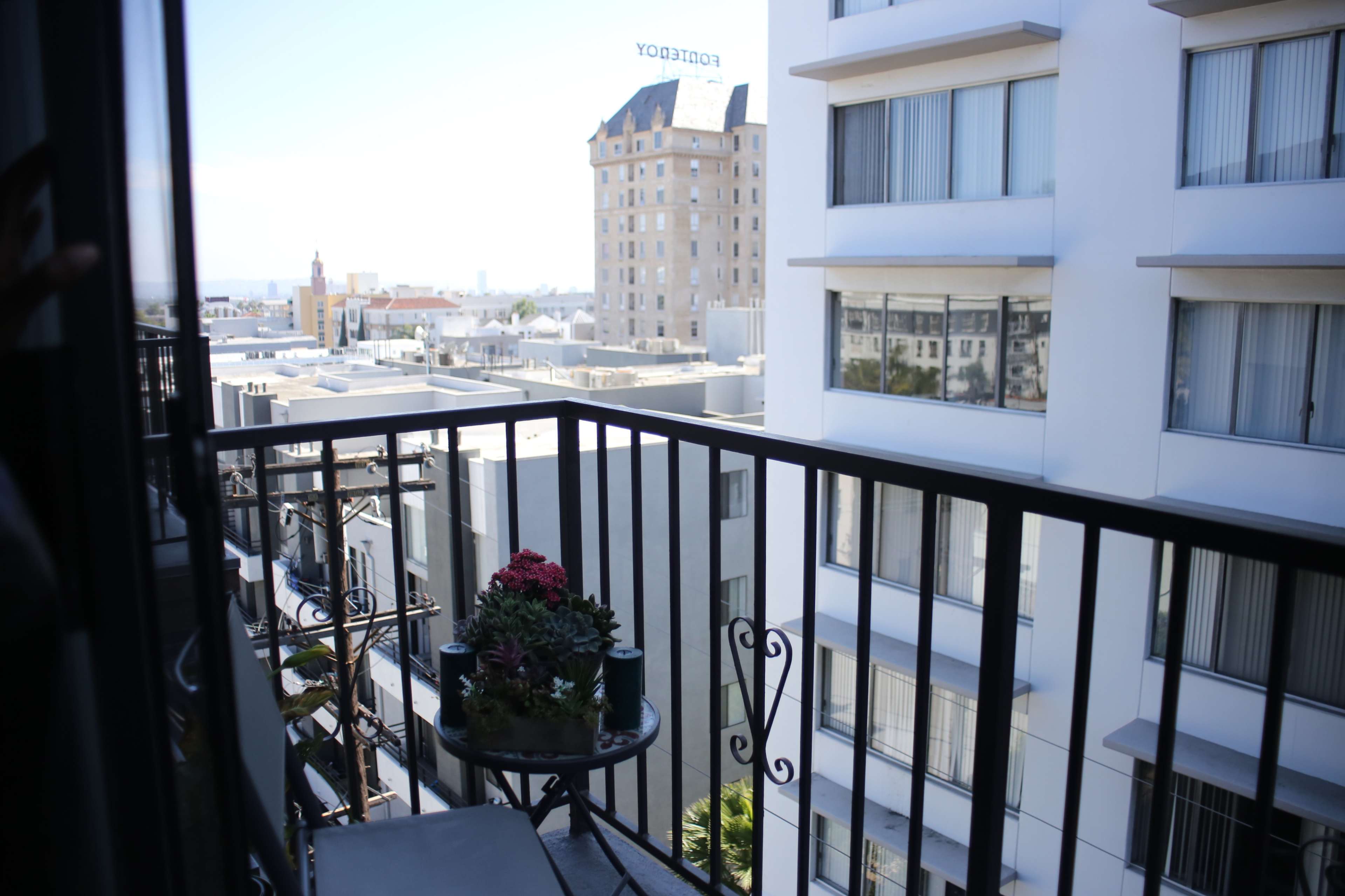 A balcony with a small table and potted plants, overlooking a cityscape with buildings in the distance.