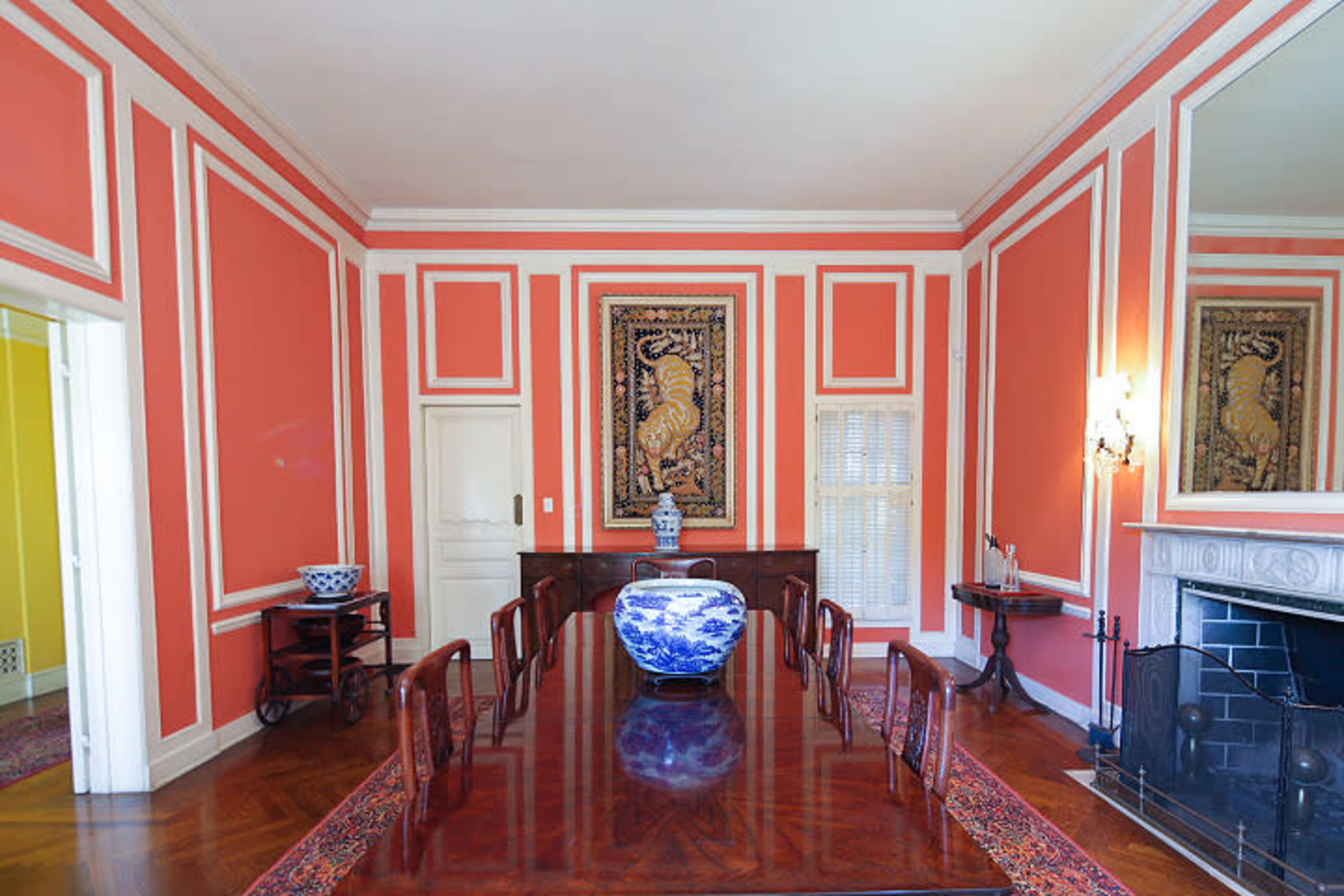 A brightly colored dining room with coral walls, a long wooden table, and a large blue and white vase as the centerpiece.
