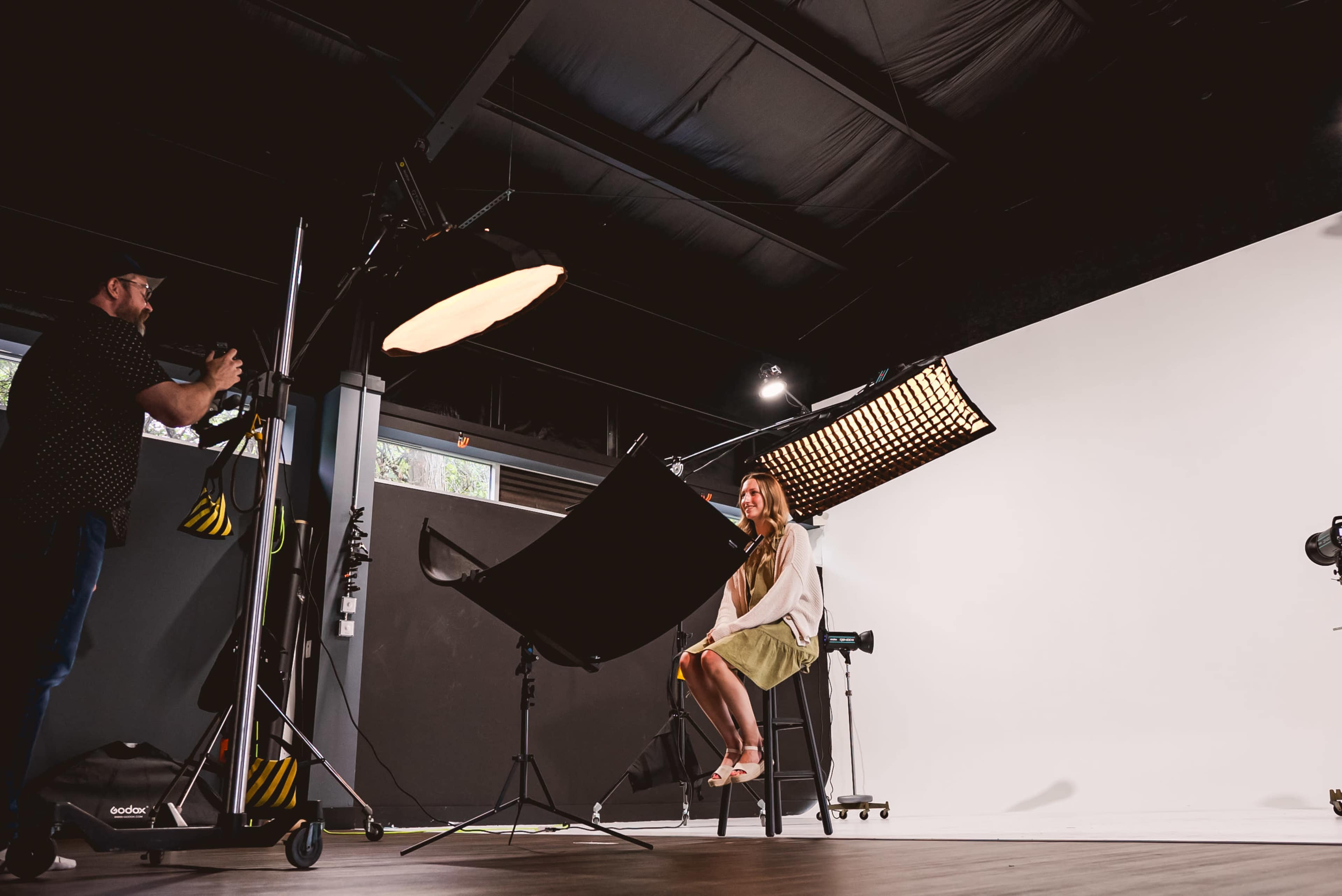 A woman sits on a stool in a photography studio while a photographer adjusts lighting equipment.