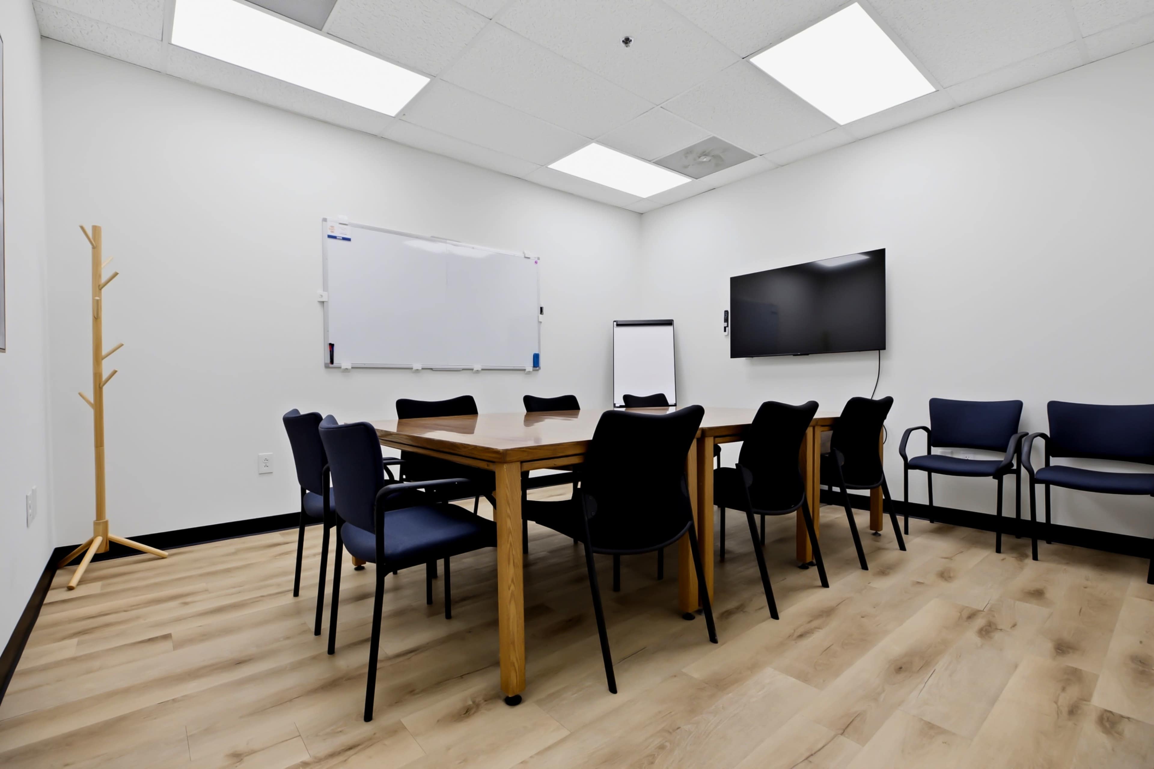 The image shows a small conference room with a wooden table surrounded by black chairs, a whiteboard on the wall, and a television mounted in one corner.