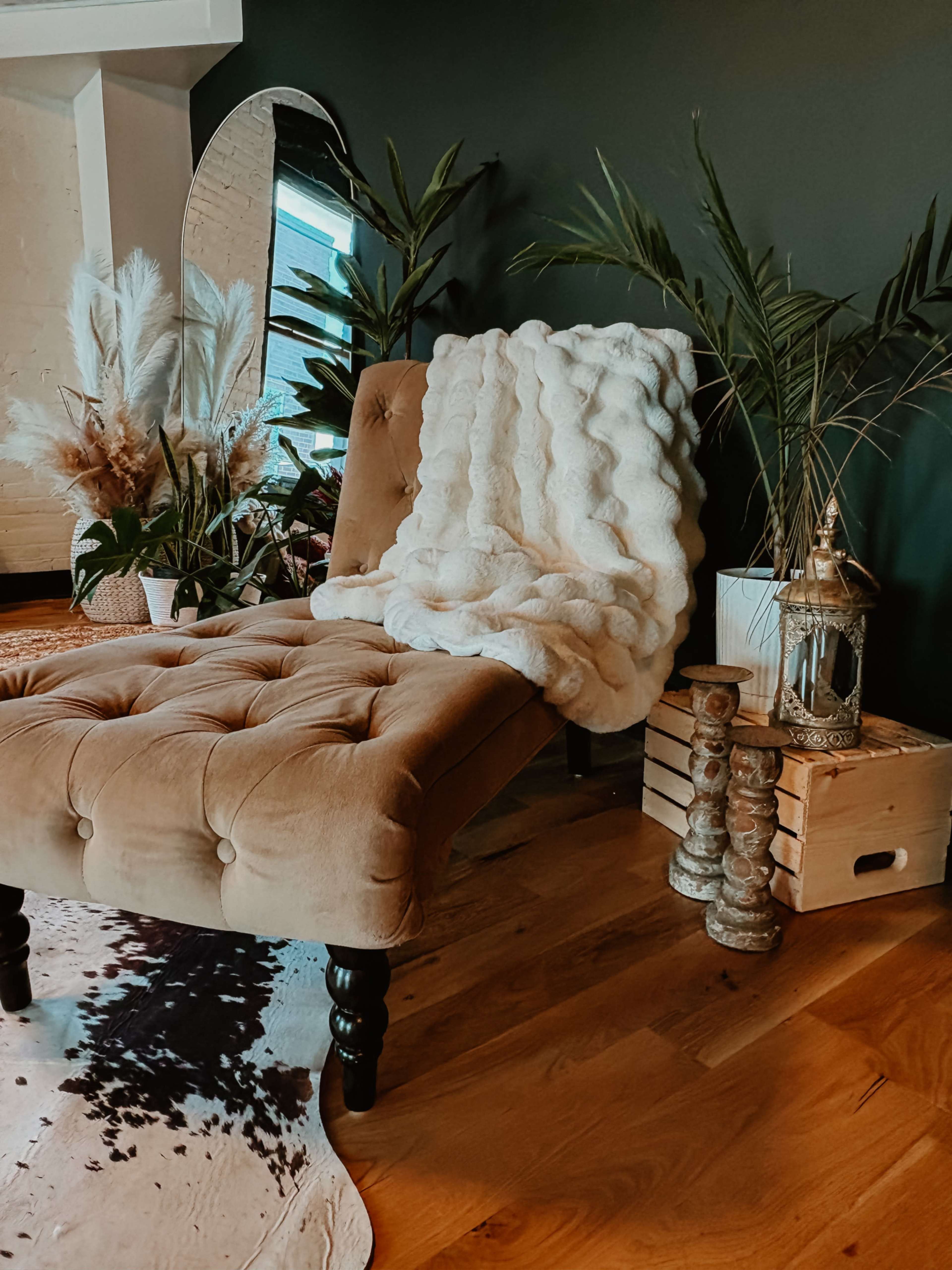 A tufted chaise lounge with a white throw is placed beside a variety of potted plants and decorative items on a wooden floor.