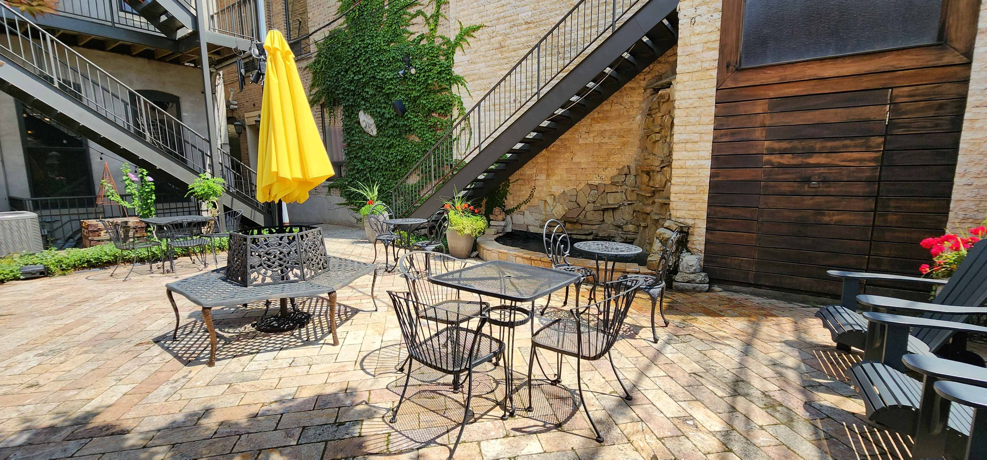 A courtyard with metal tables and chairs, a yellow umbrella, and stone walls adorned with greenery.