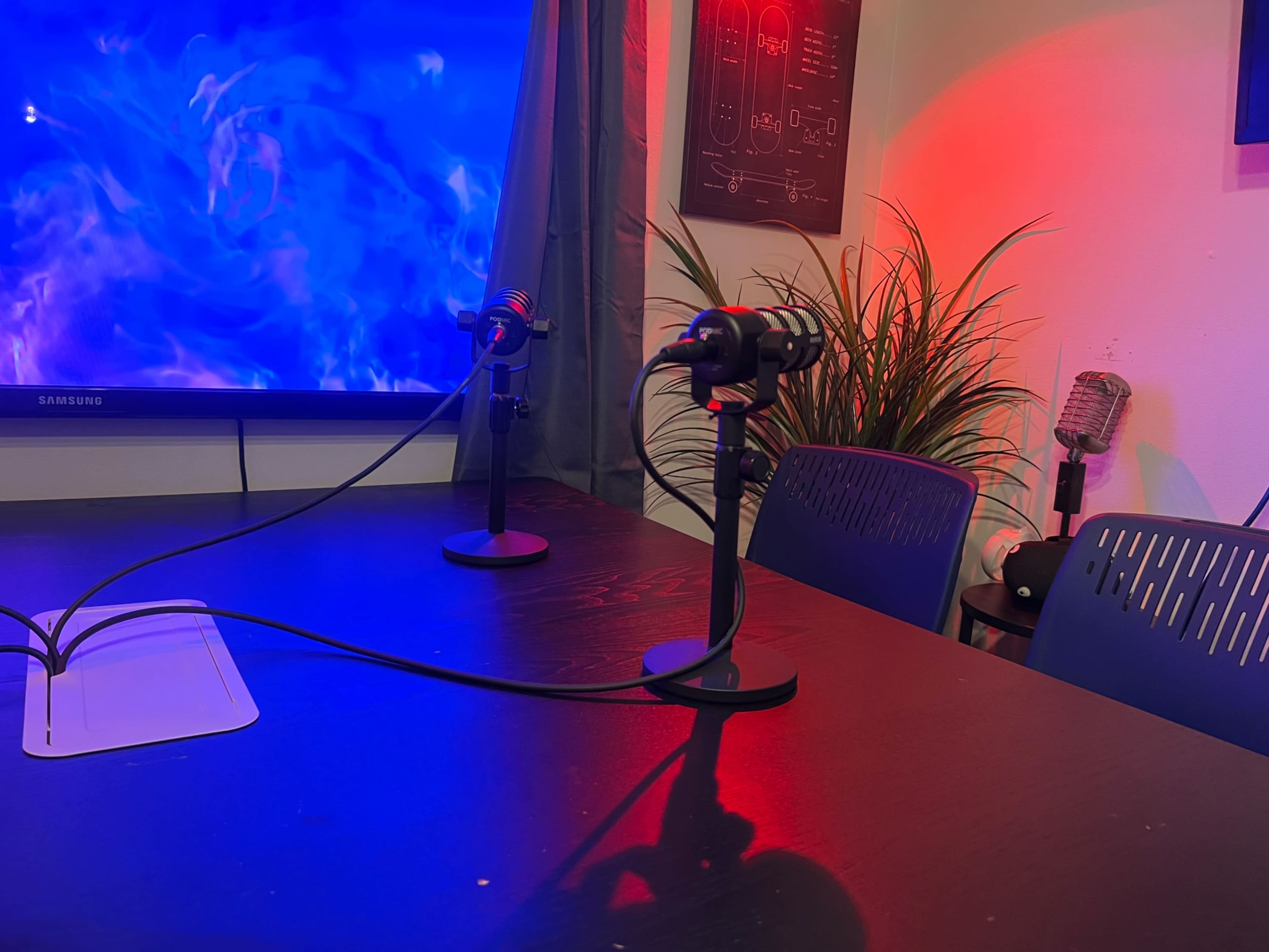 A table with two microphones, a large screen displaying abstract visuals in the background, and a plant on the side.