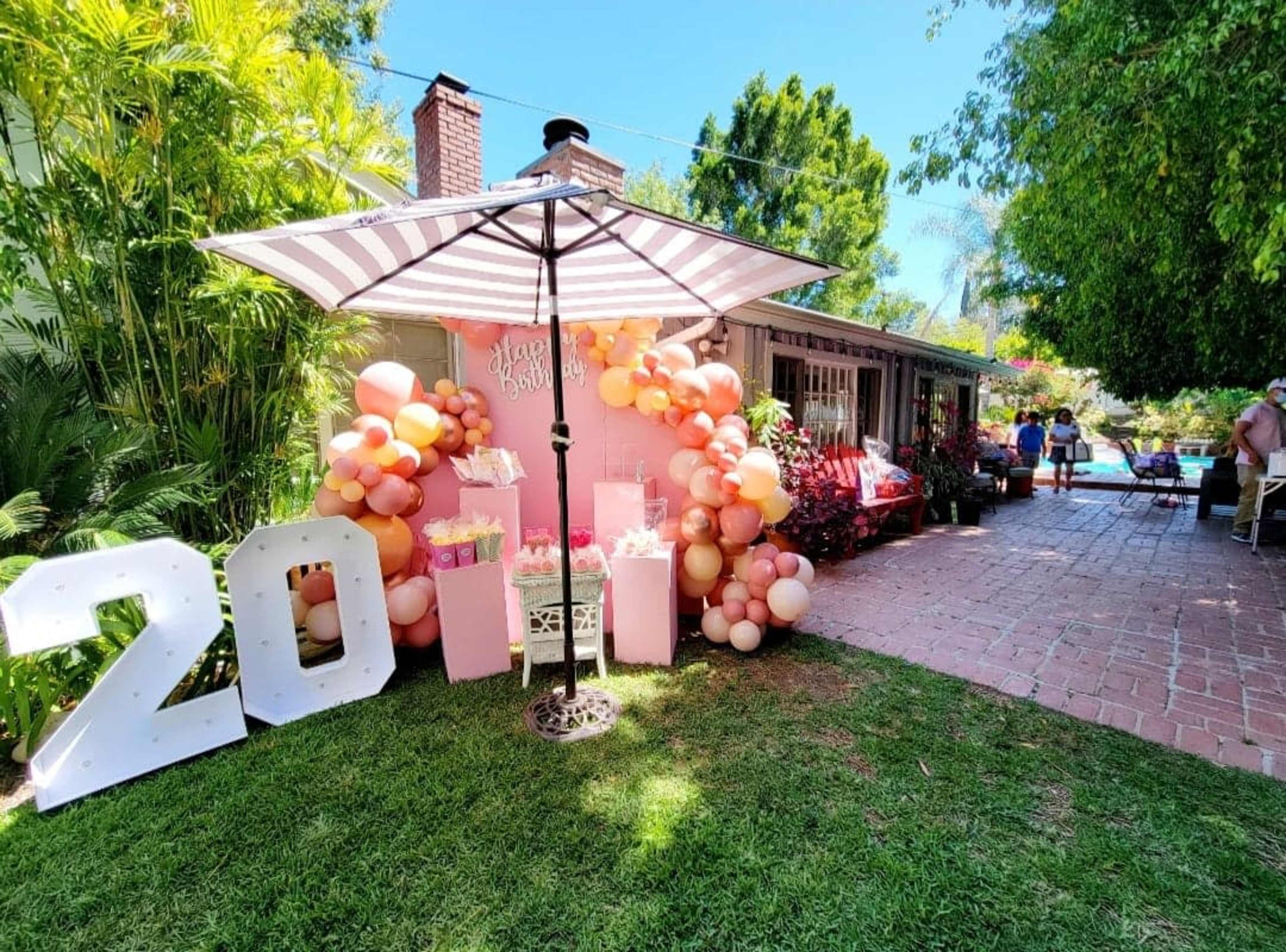 A birthday celebration scene features a large "20" display next to a festive balloon arrangement and gifts under a striped umbrella, with guests mingling near a pool in the background.