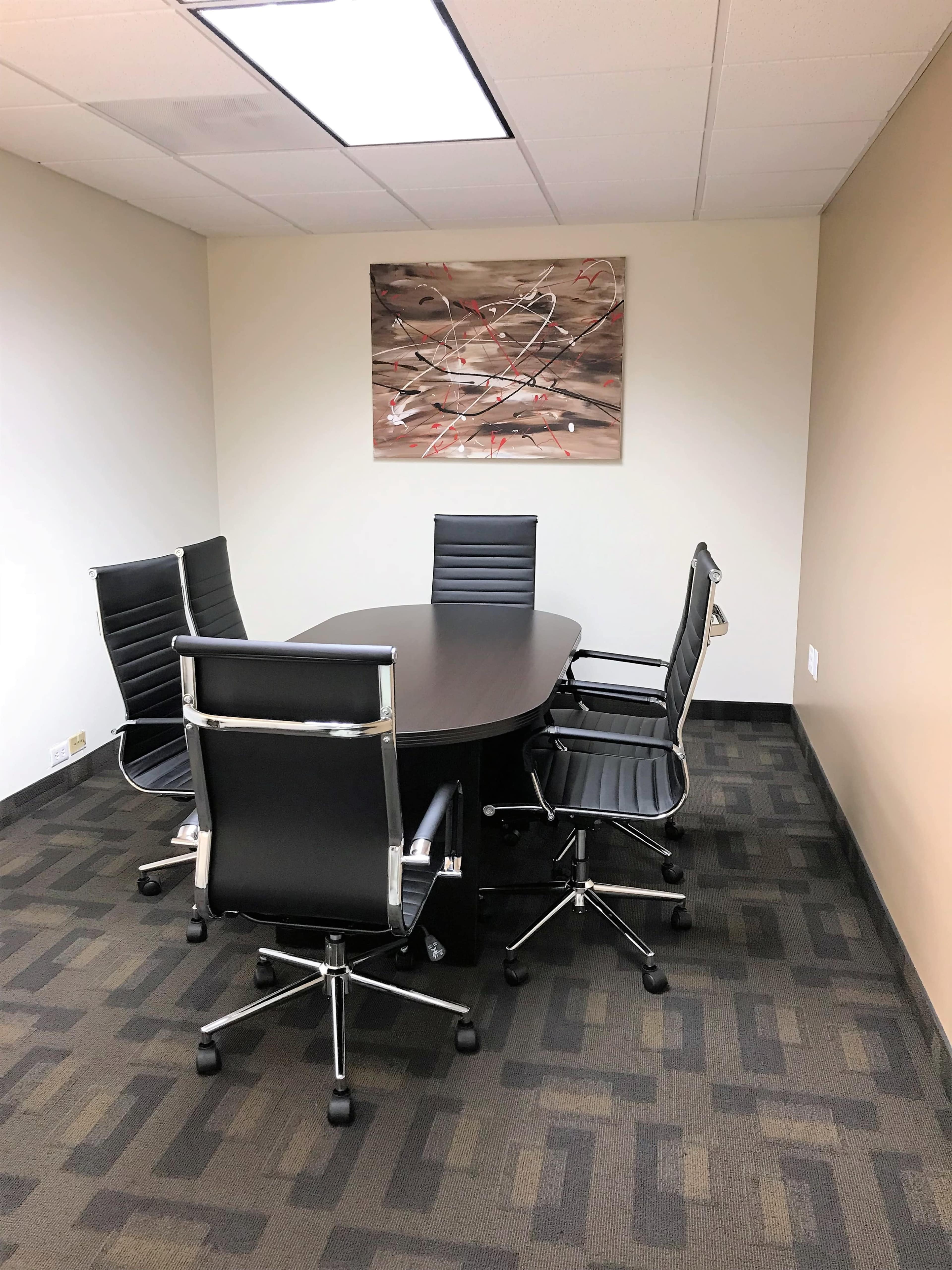 A conference room features a large oval table surrounded by four black office chairs and a piece of abstract art on the wall.