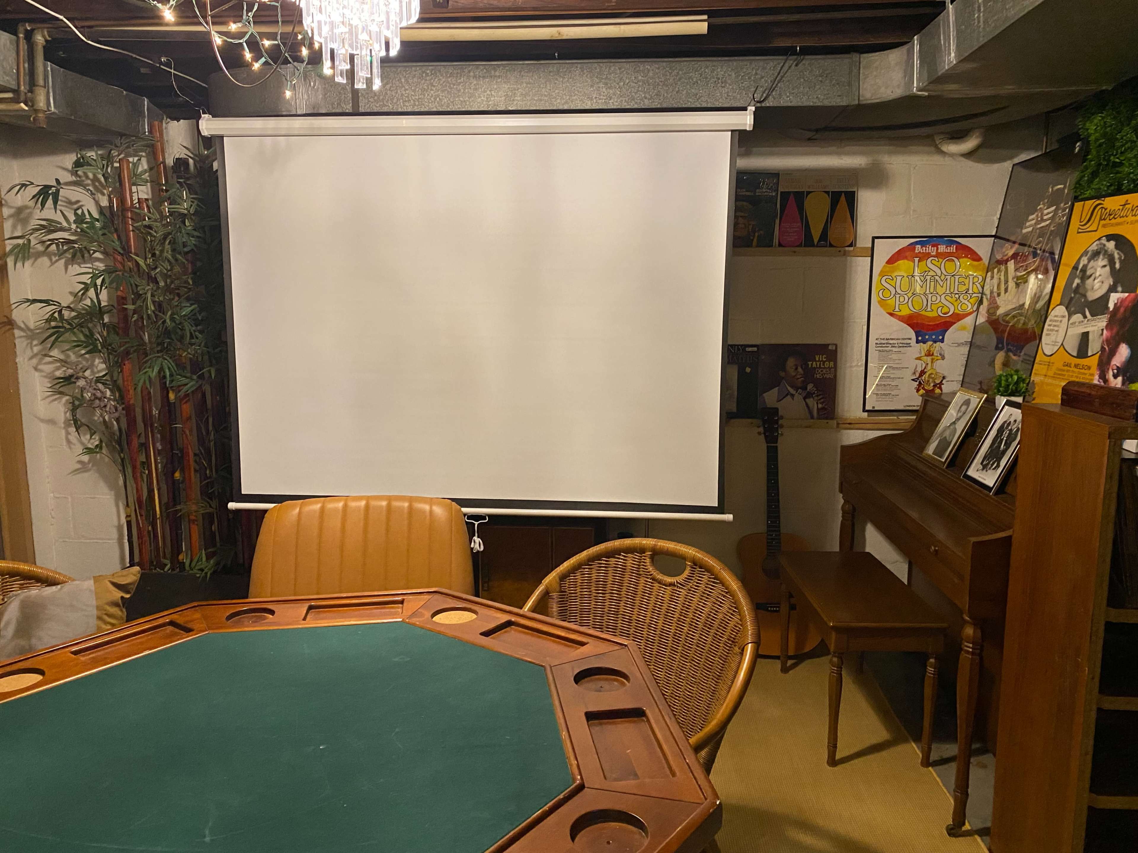 The image shows a basement room with a round table, a projector screen, a wooden piano, and a few seating arrangements, including wicker chairs.