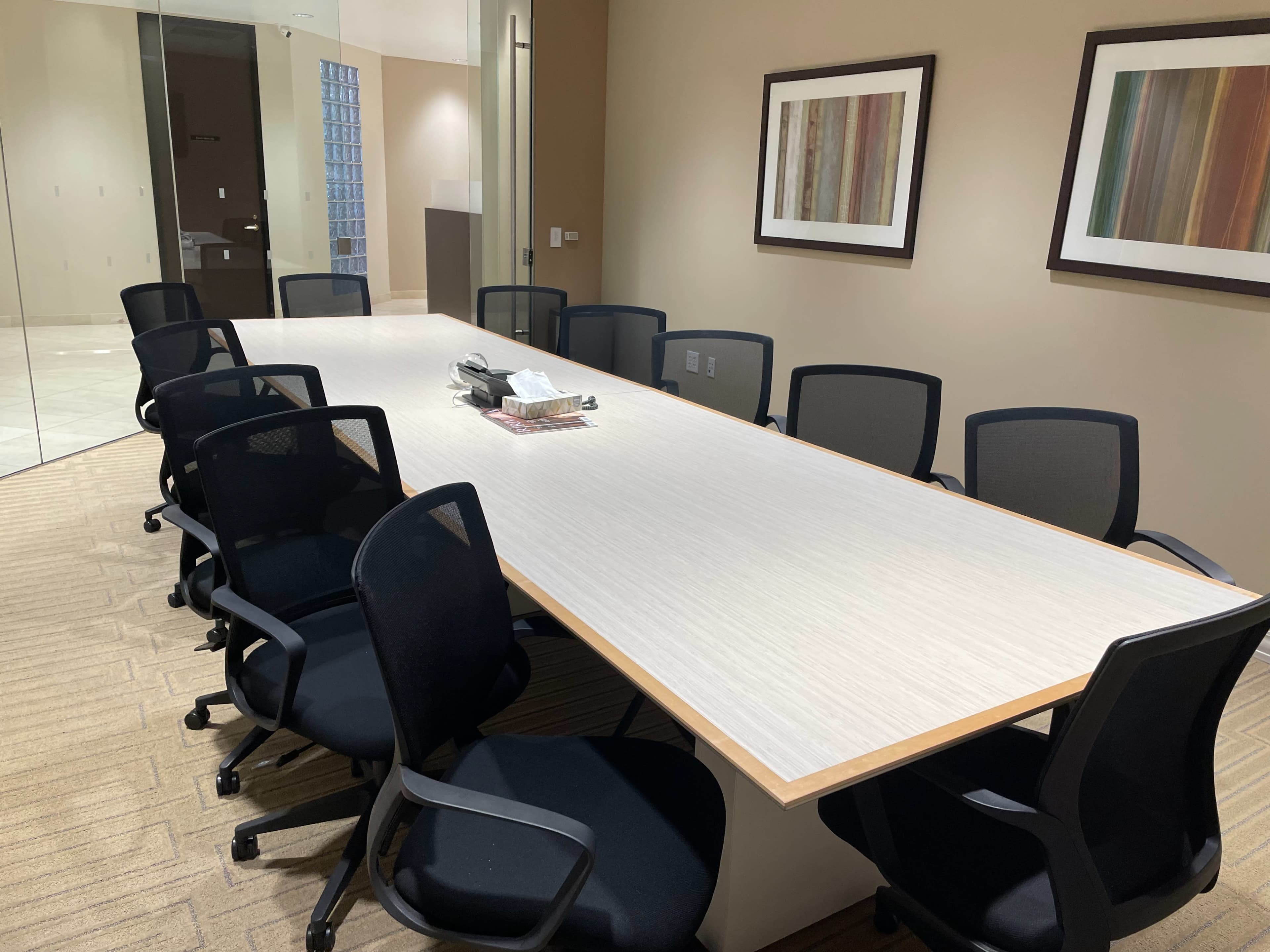A long conference table is surrounded by black office chairs in a well-lit meeting room, flanked by two framed artworks on the walls.