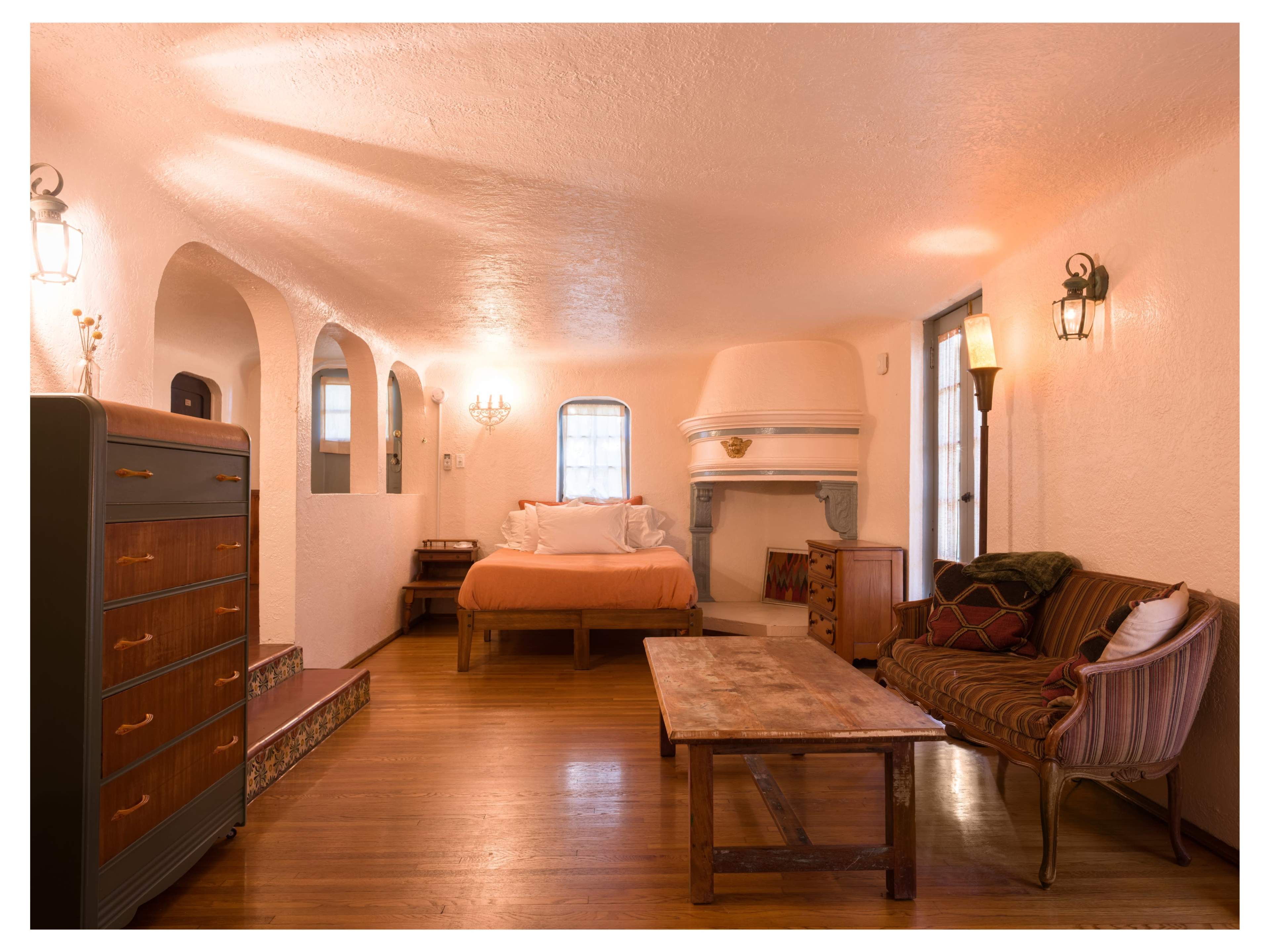 The image shows a cozy, light-colored room featuring a bed, a fireplace, a wooden table, and seating arrangements, with arched doorways and warm lighting.