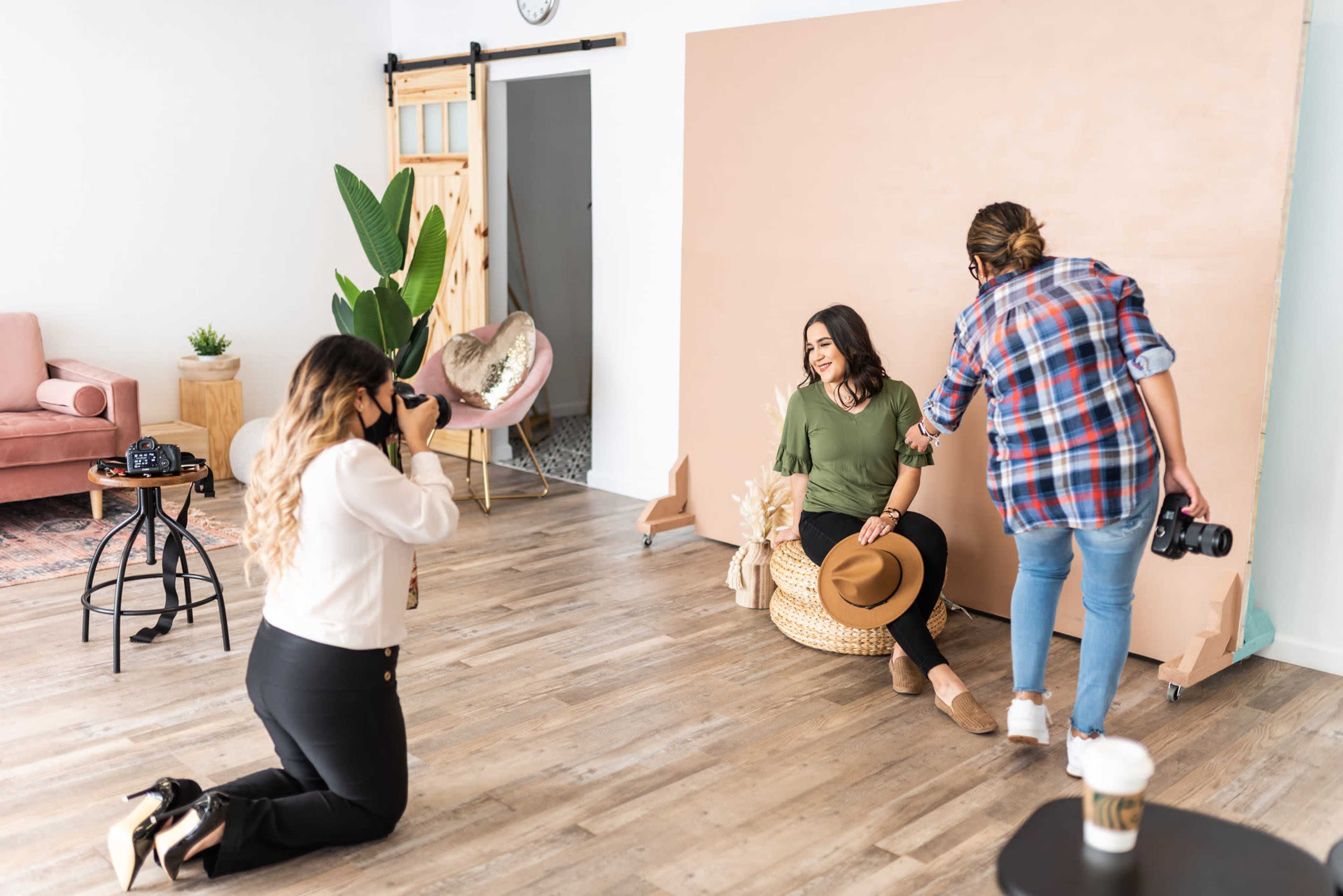 A photoshoot takes place in a studio with a model sitting on a chair, while a photographer captures the moment and a stylist adjusts the scene.