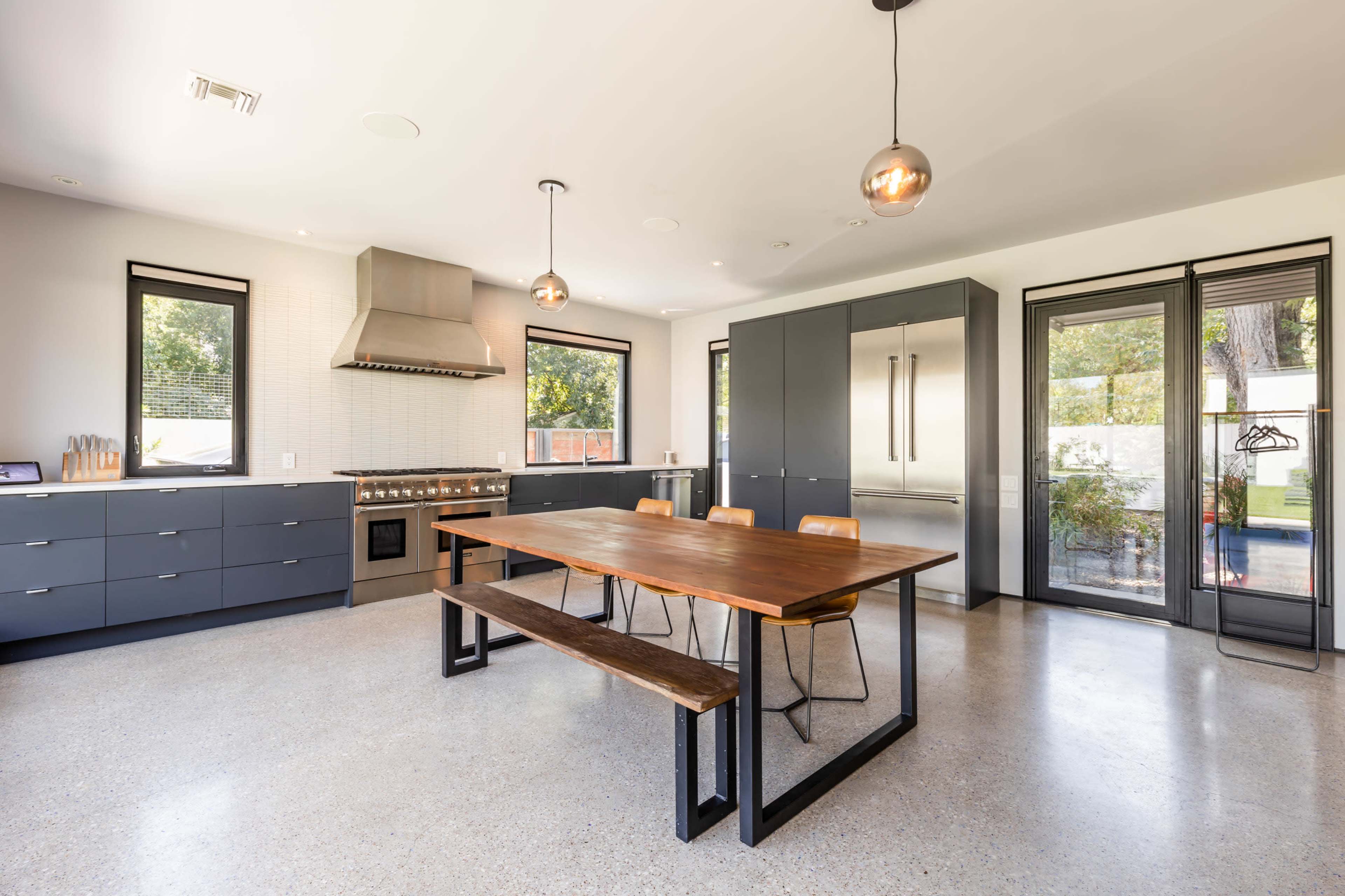 A modern kitchen features a central wooden table surrounded by chairs, stainless steel appliances, and dark cabinetry.