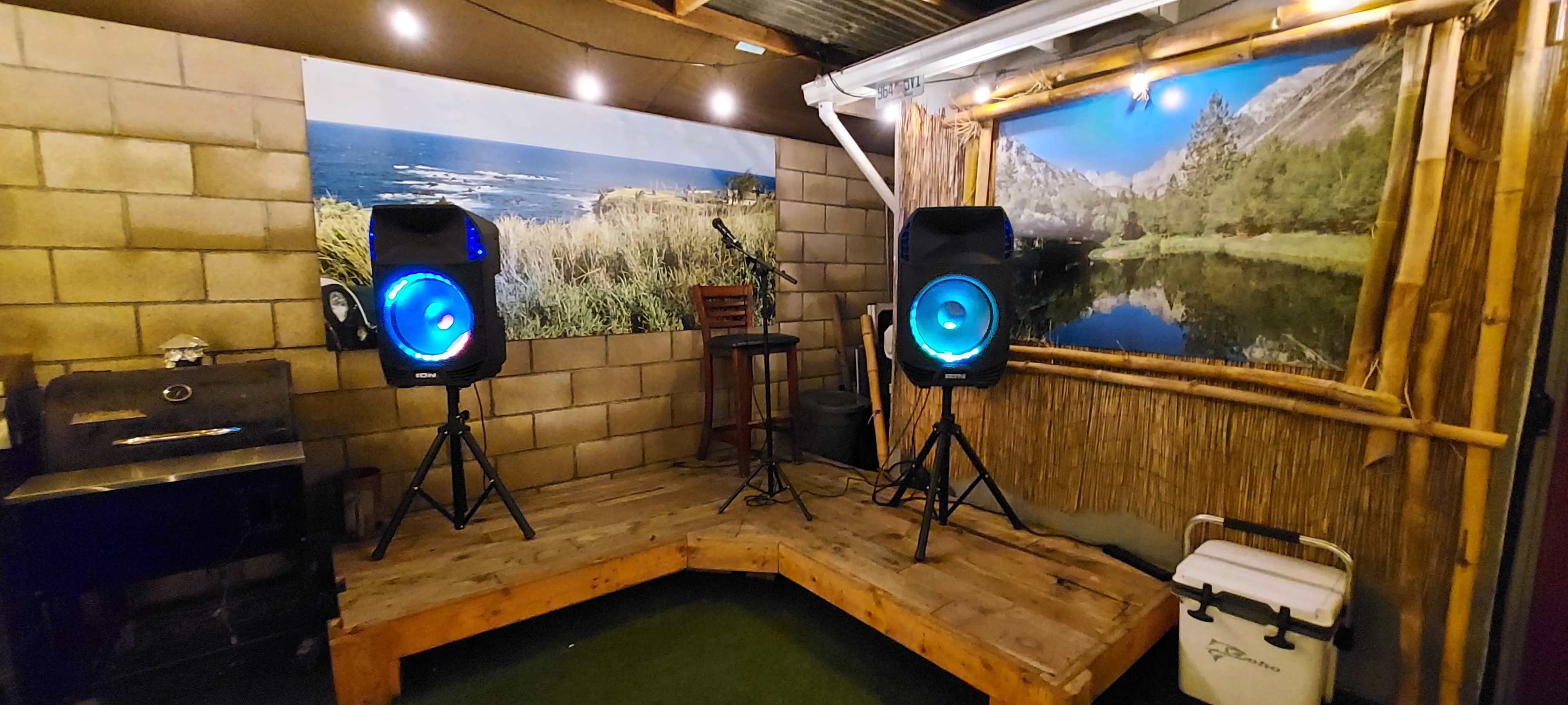 A small performance stage with two speakers, a microphone stand, and a stool, set against a backdrop of a scenic landscape.