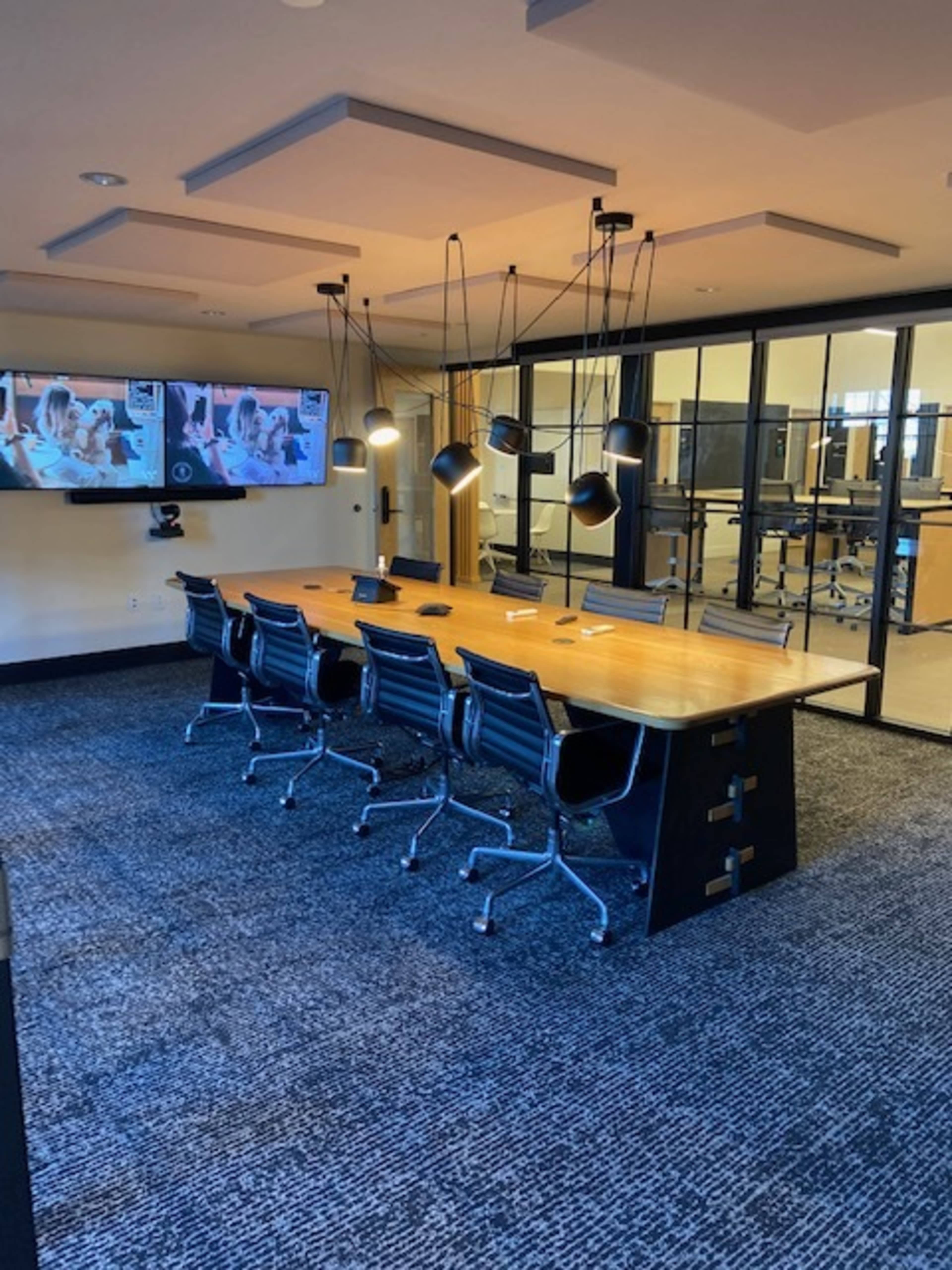 A modern conference room with a large wooden table, six black ergonomic chairs, and a wall-mounted screen displaying a video conference setup.