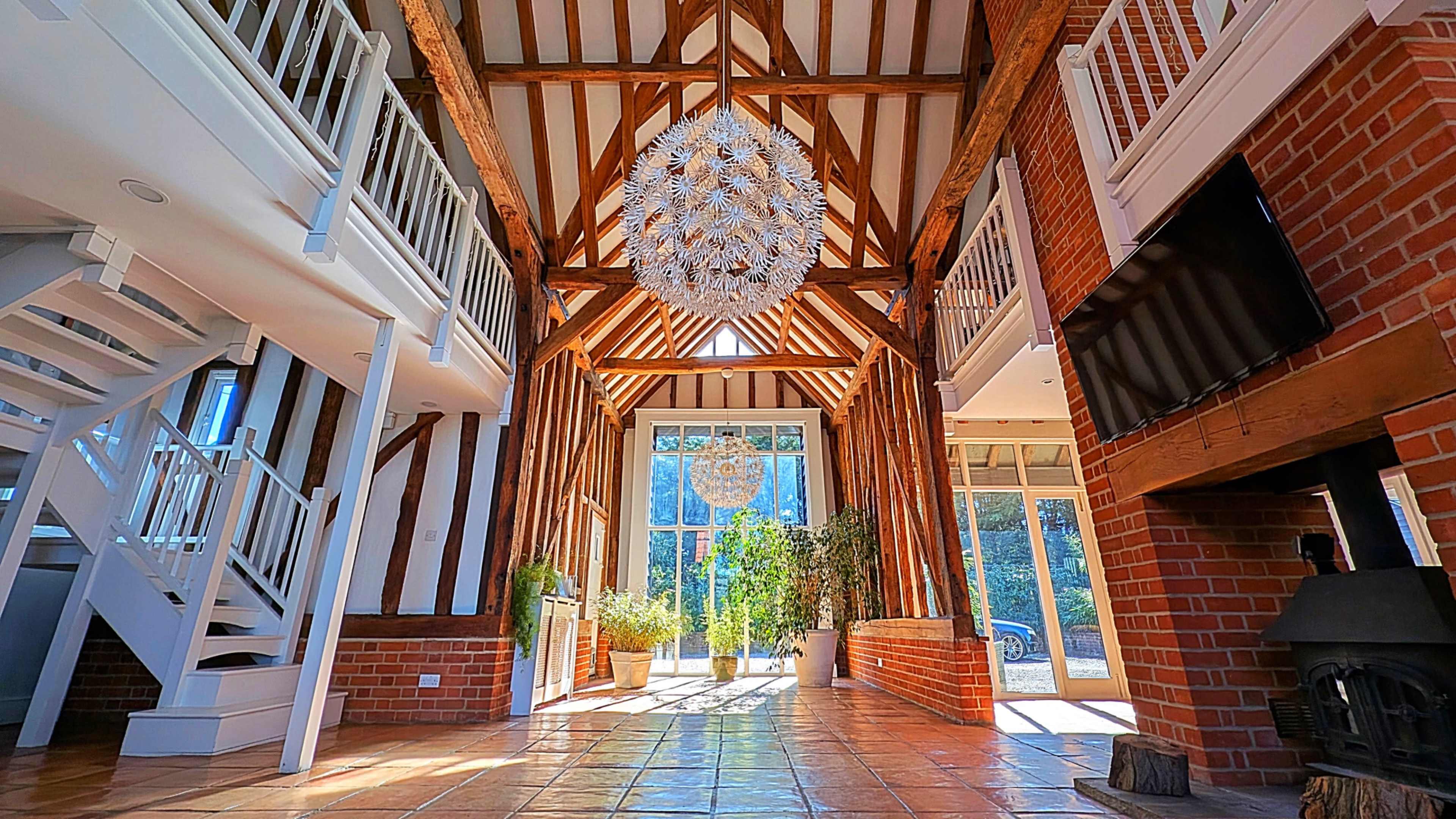 The image shows a spacious interior with exposed wooden beams, a large chandelier, and a staircase leading to an upper level, flanked by large windows.