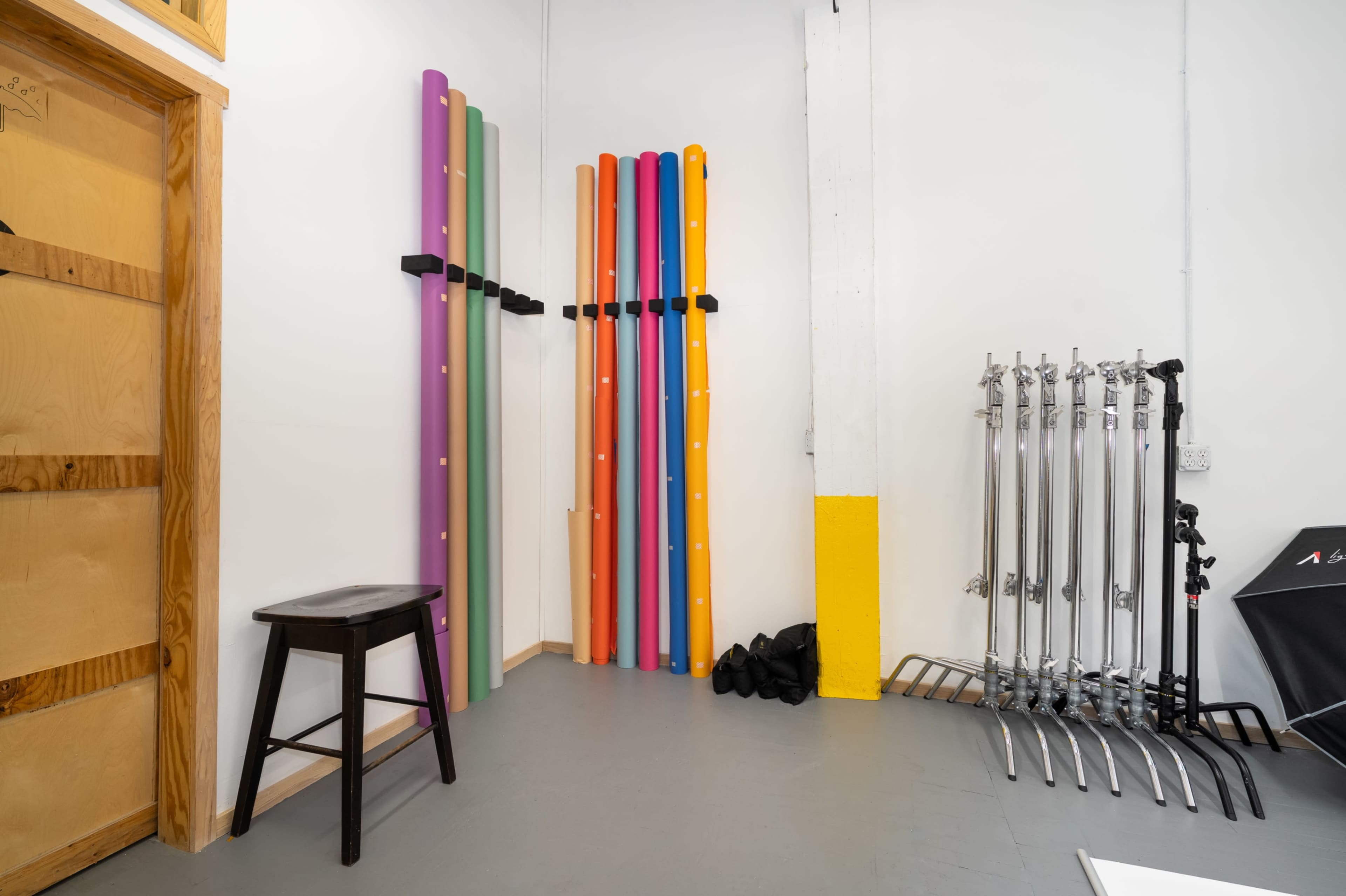 The image shows a brightly colored wall-mounted storage of rolls next to a metal rack with several tubular structures and a black stool in a spacious room with gray flooring.